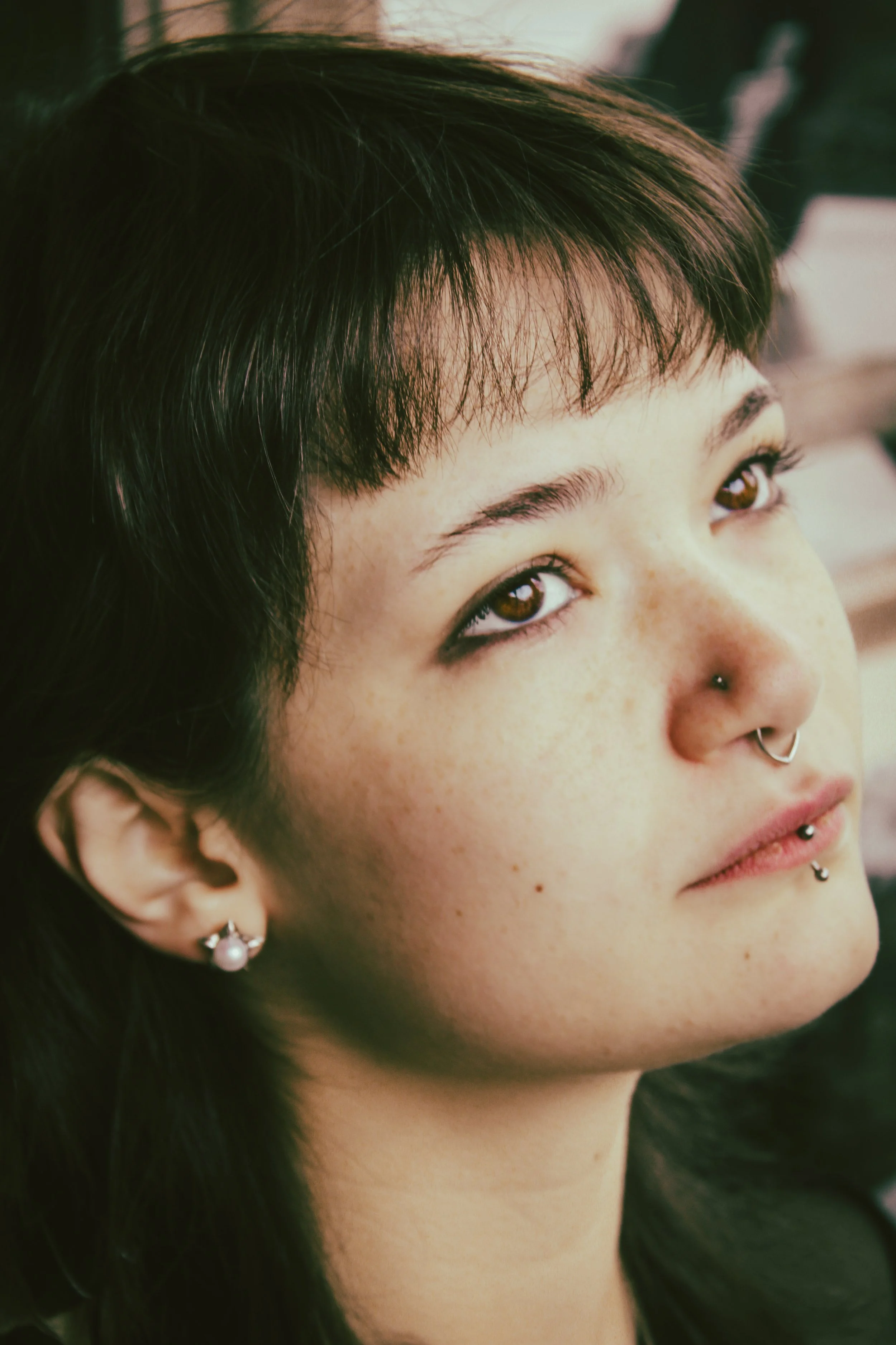 A close-up of a young woman with dark hair, brown eyes, and multiple facial piercings, including a nose ring, a labret piercing, and a septum piercing.
