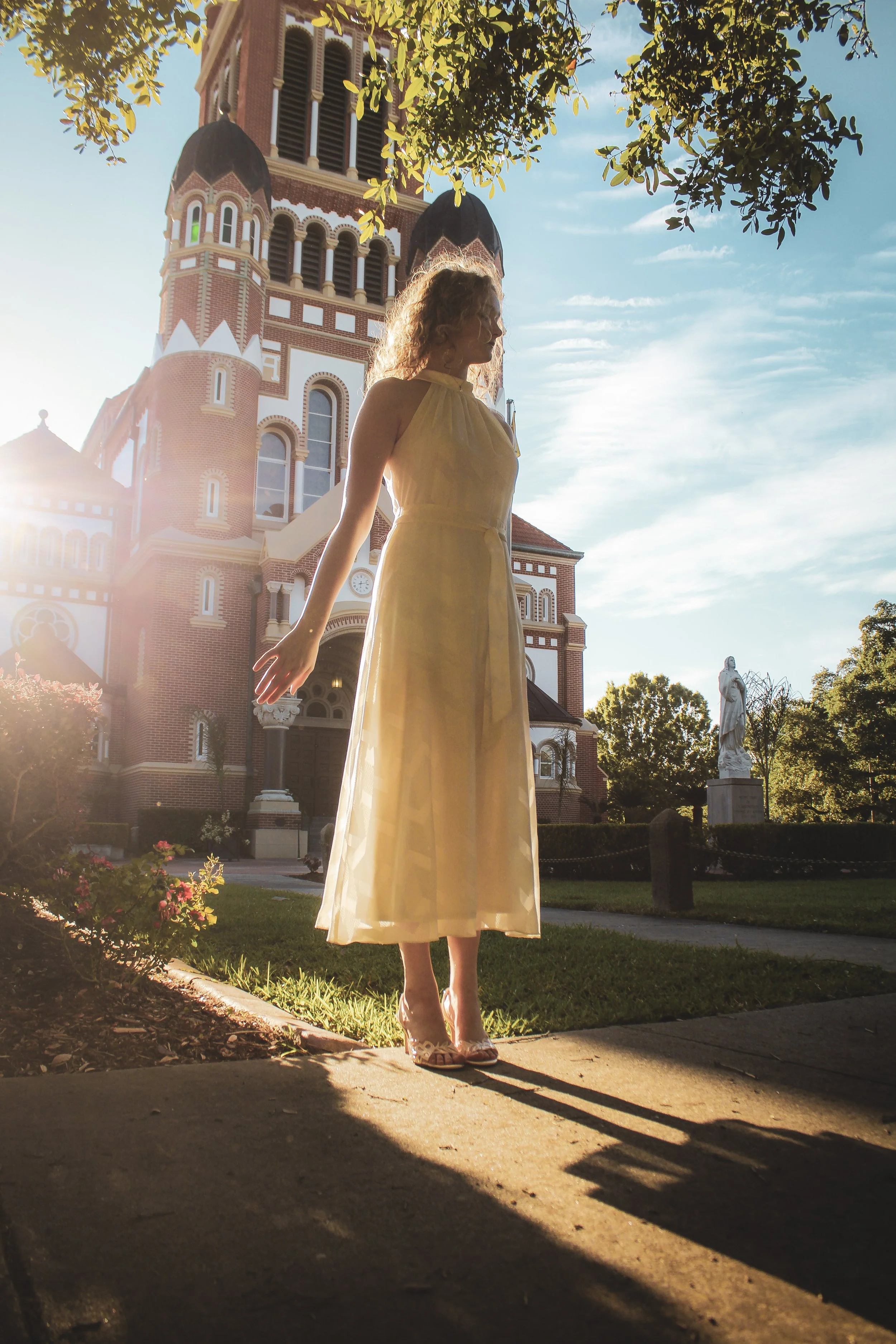 A woman in a yellow dress standing on a sidewalk in front of a large brick church with a tall steeple, illuminated by the warm glow of the setting sun, with trees and statues surrounding the scene.