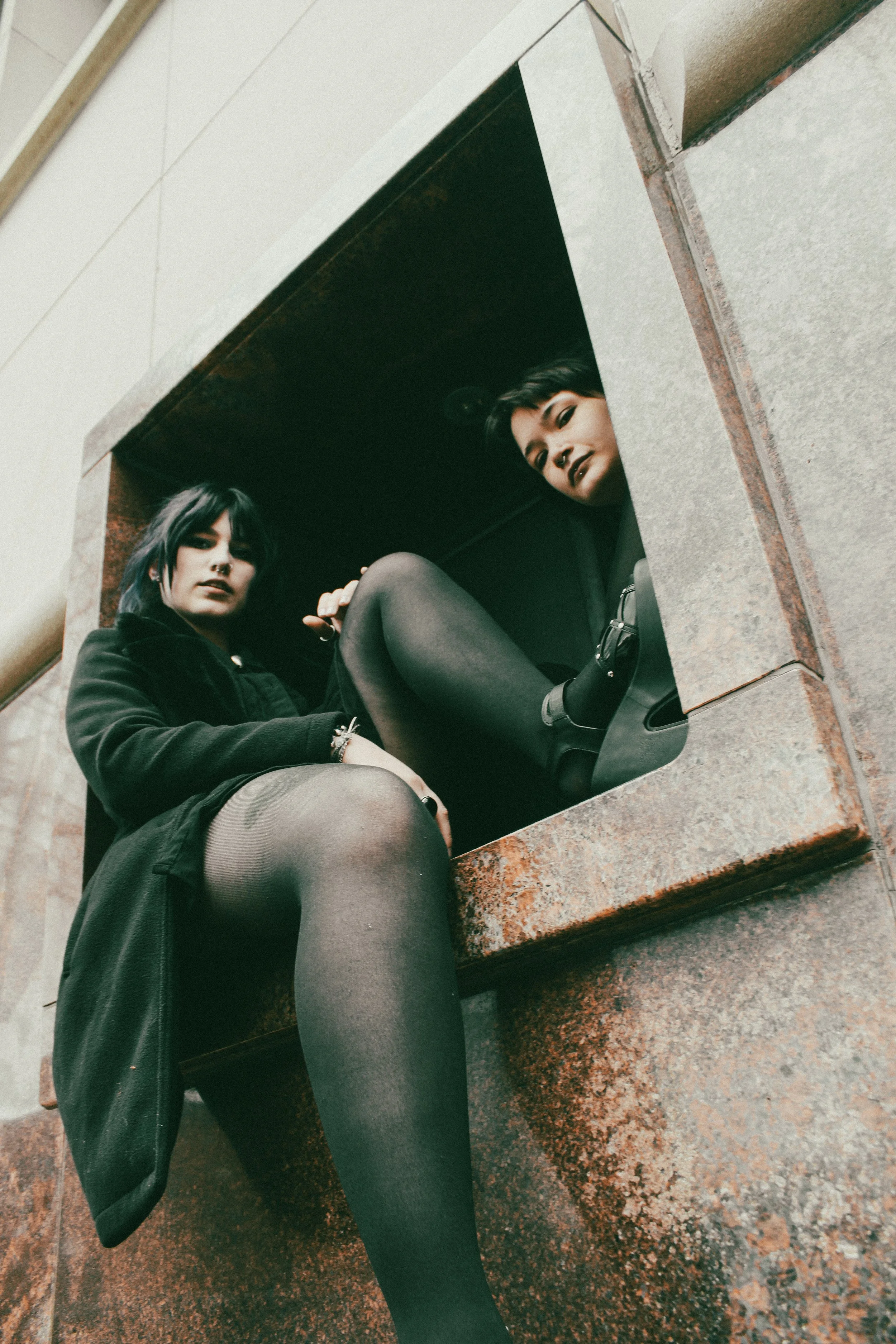 Two women with dark hair sitting inside a small window or opening in a brick or concrete structure, looking down at the camera.