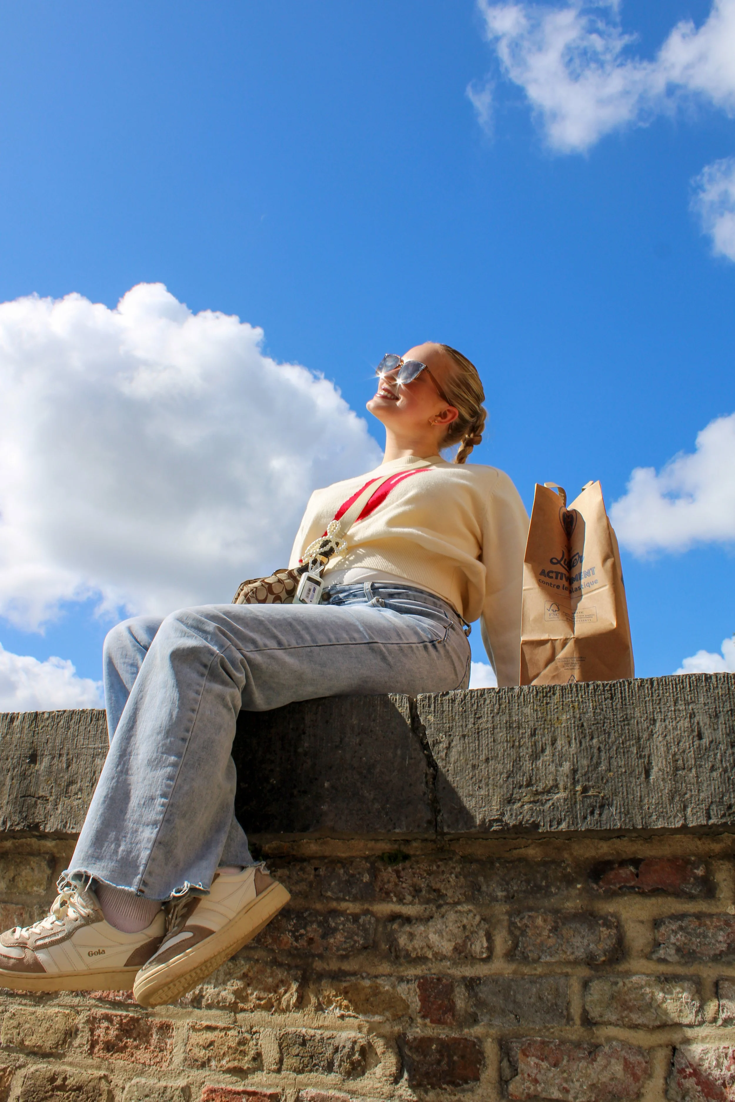 A woman sitting on a brick wall, smiling and enjoying the sunny weather, with a bright blue sky and clouds in the background, wearing sunglasses, a sweater, jeans, and sneakers, with shopping bags nearby.