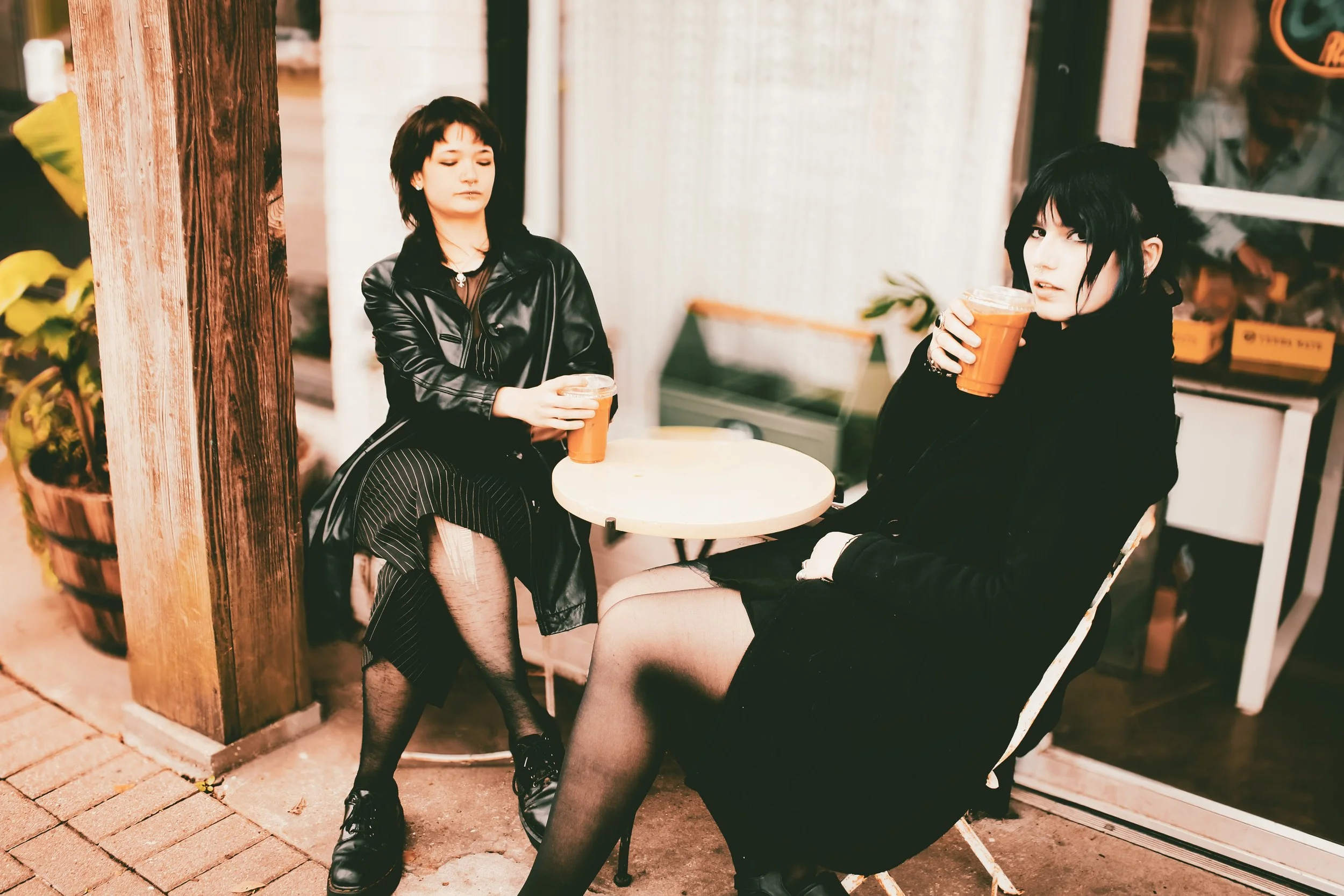 Two women sitting outside a cafe at a small round table, each holding a drink in a plastic cup with an orange liquid, dressed in dark clothing and black tights, with a wooden pillar and a plant nearby.