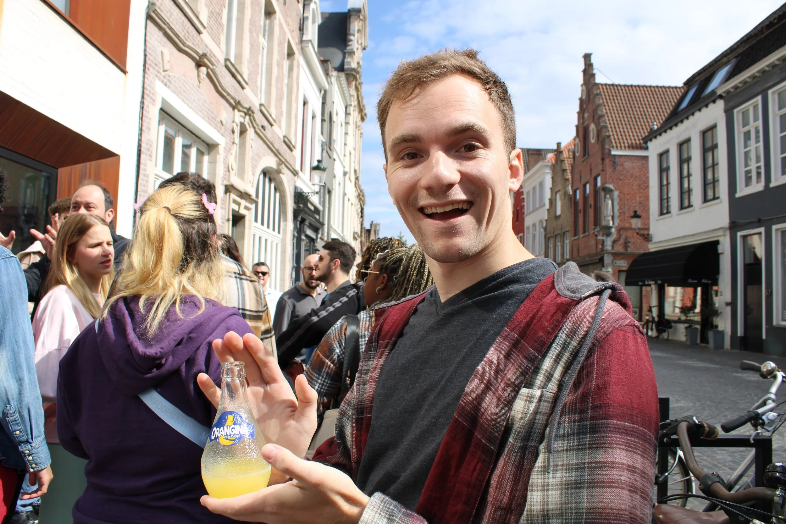 A smiling young man holding a bottle of Orangina soda outdoors among a crowd of people on a city street.