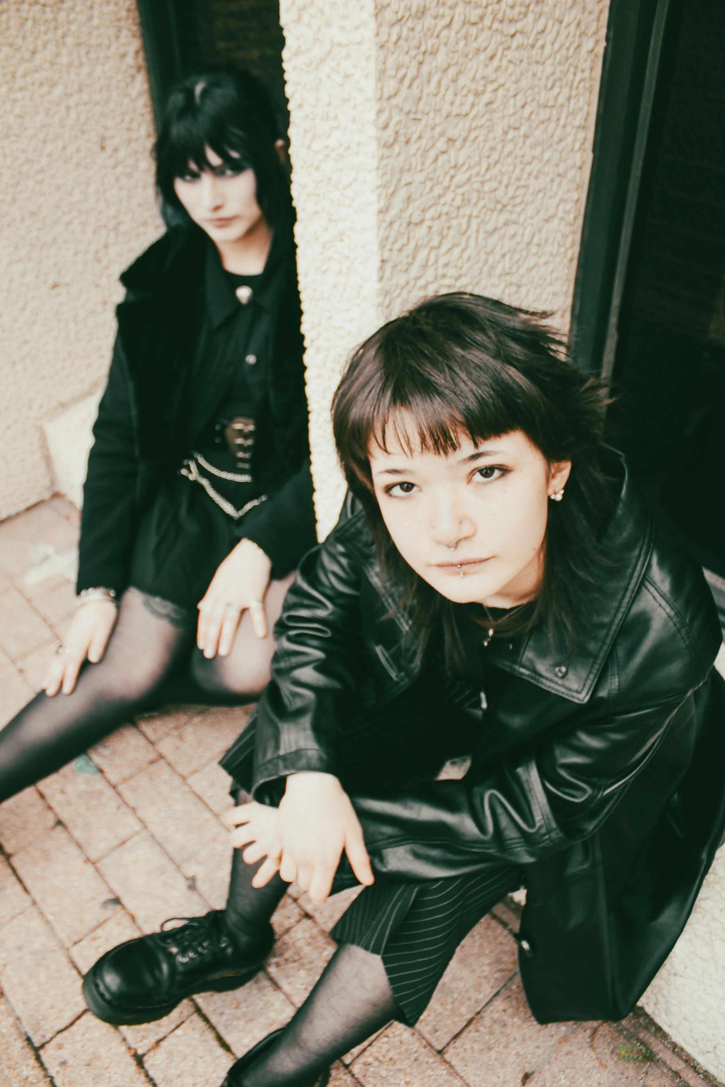 Two young women with dark hair styled in emo fashion, wearing black clothing, sitting on brick pavement near a beige stucco wall, looking at the camera.