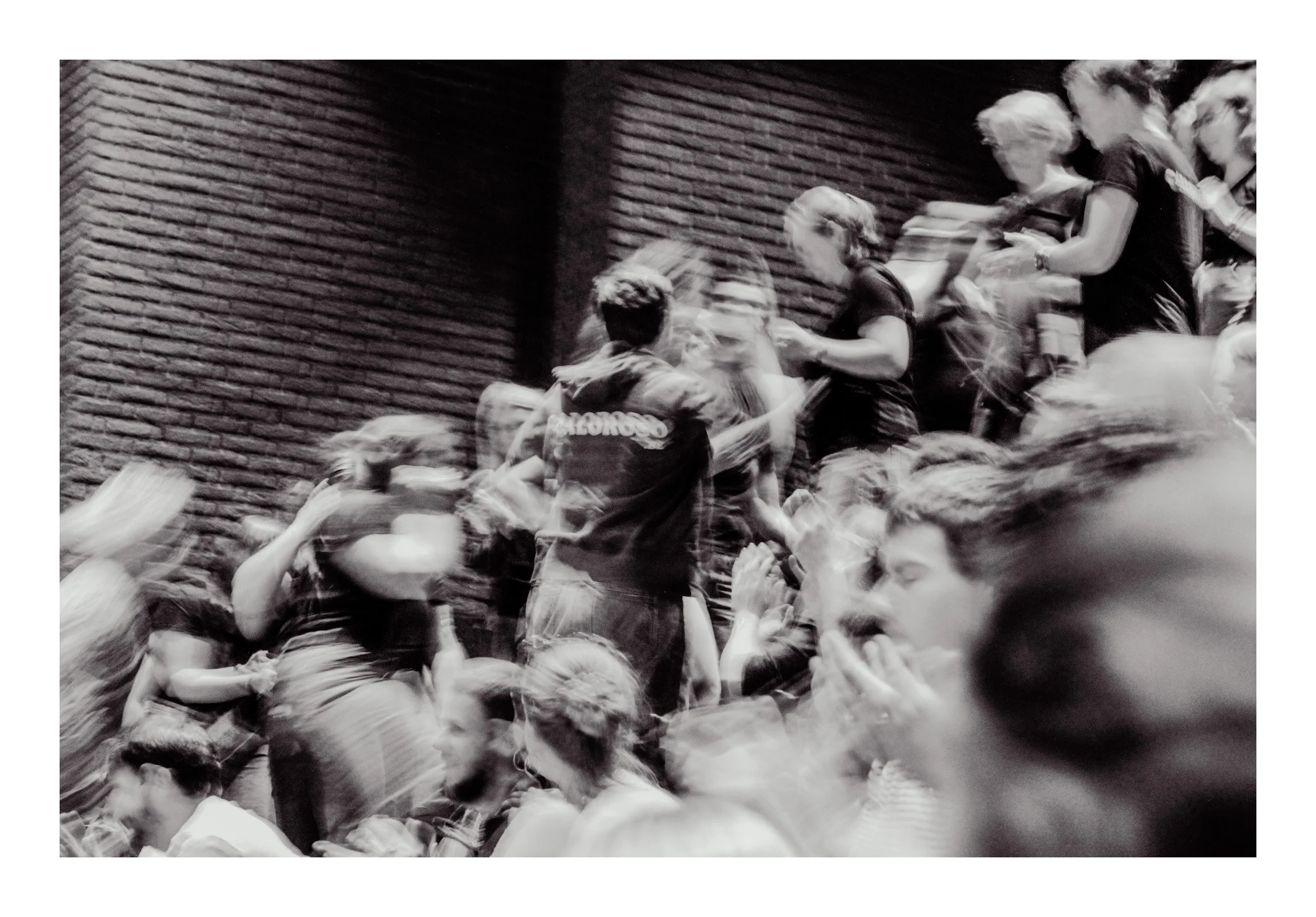 Group of people in a crowded, dynamic scene, with some individuals standing and others seated, against a brick wall background, captured in black and white with motion blur.