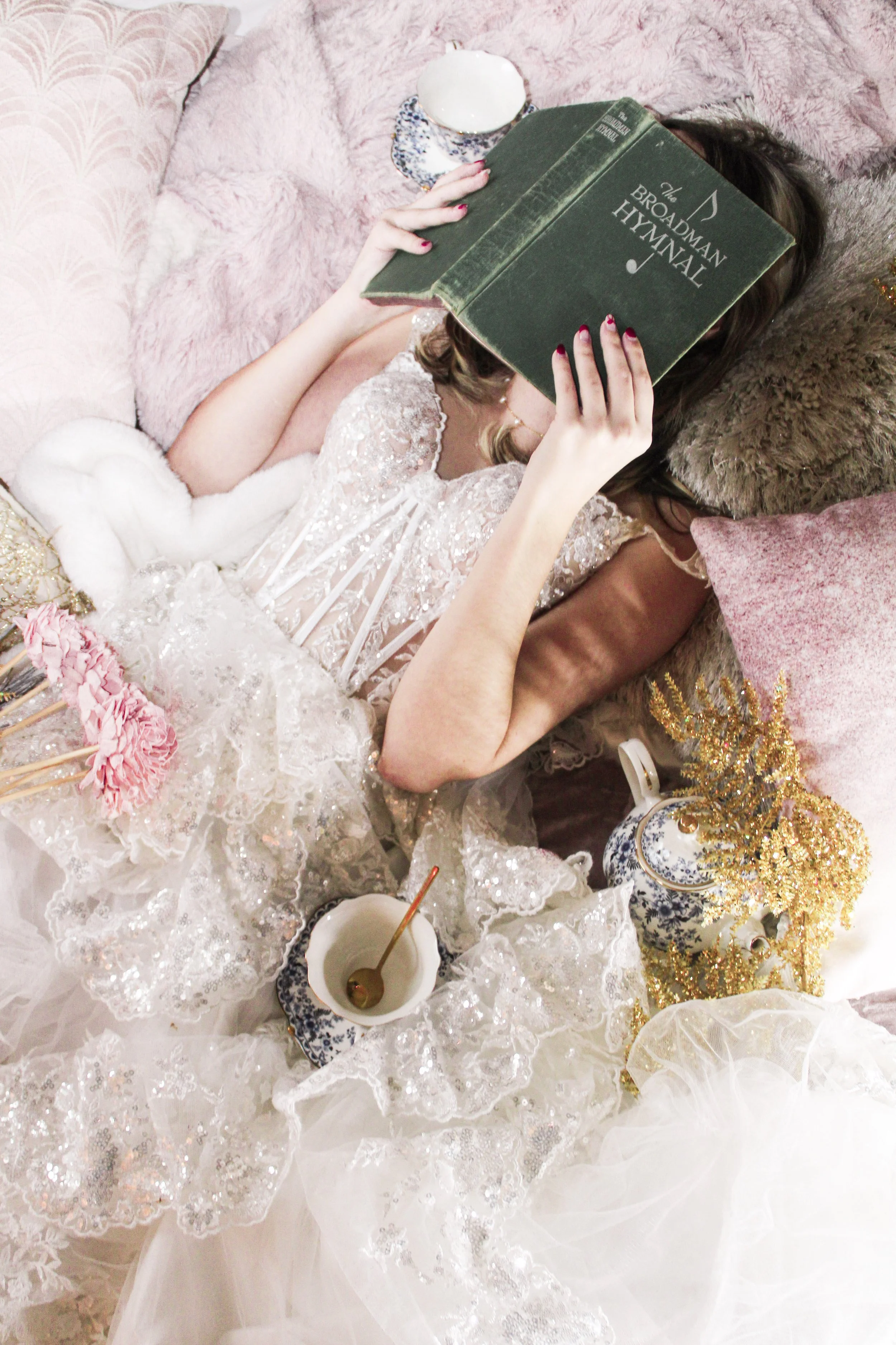 A woman in a white lace dress lies on a bed surrounded by pink and gold pillows, reading a large green book titled 'The Broadman Hymnal'.