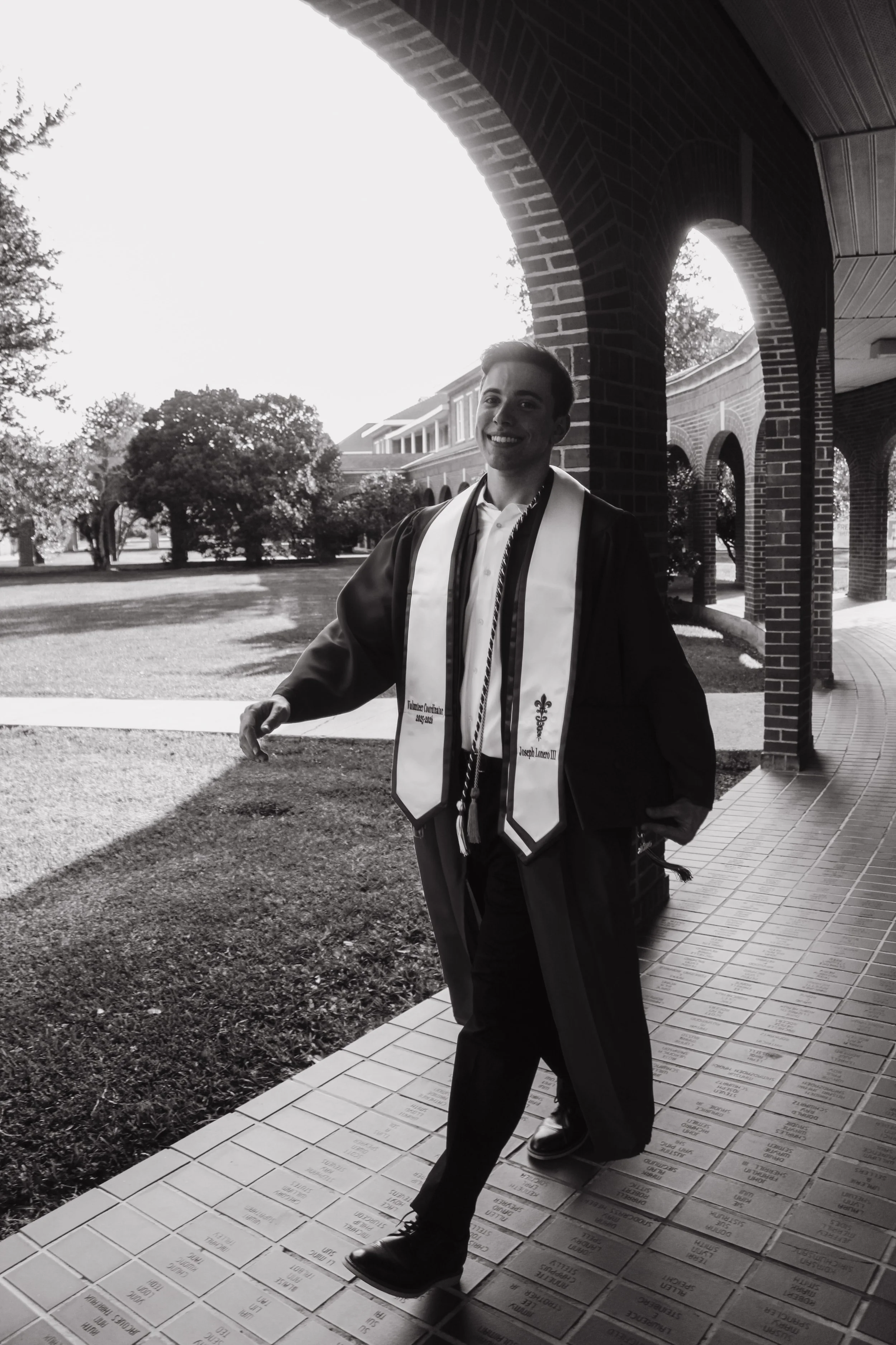 A young man wearing graduation attire, including a gown and sash, smiling and walking on engraved bricks outside a brick building with arches.