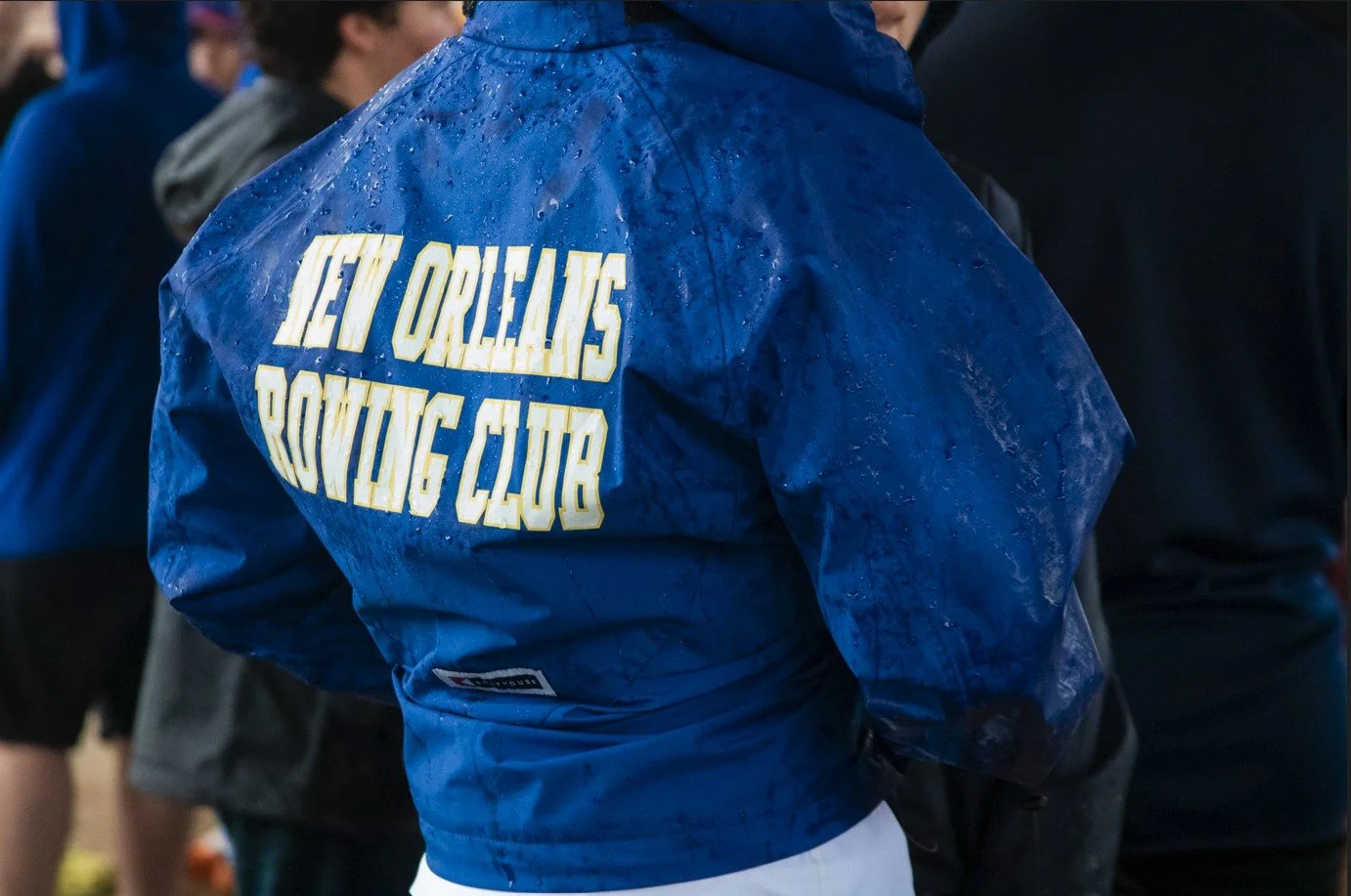 Person wearing a blue jacket with water droplets, with yellow text that reads 'New Orleans Boxing Club' on the back, among other people.