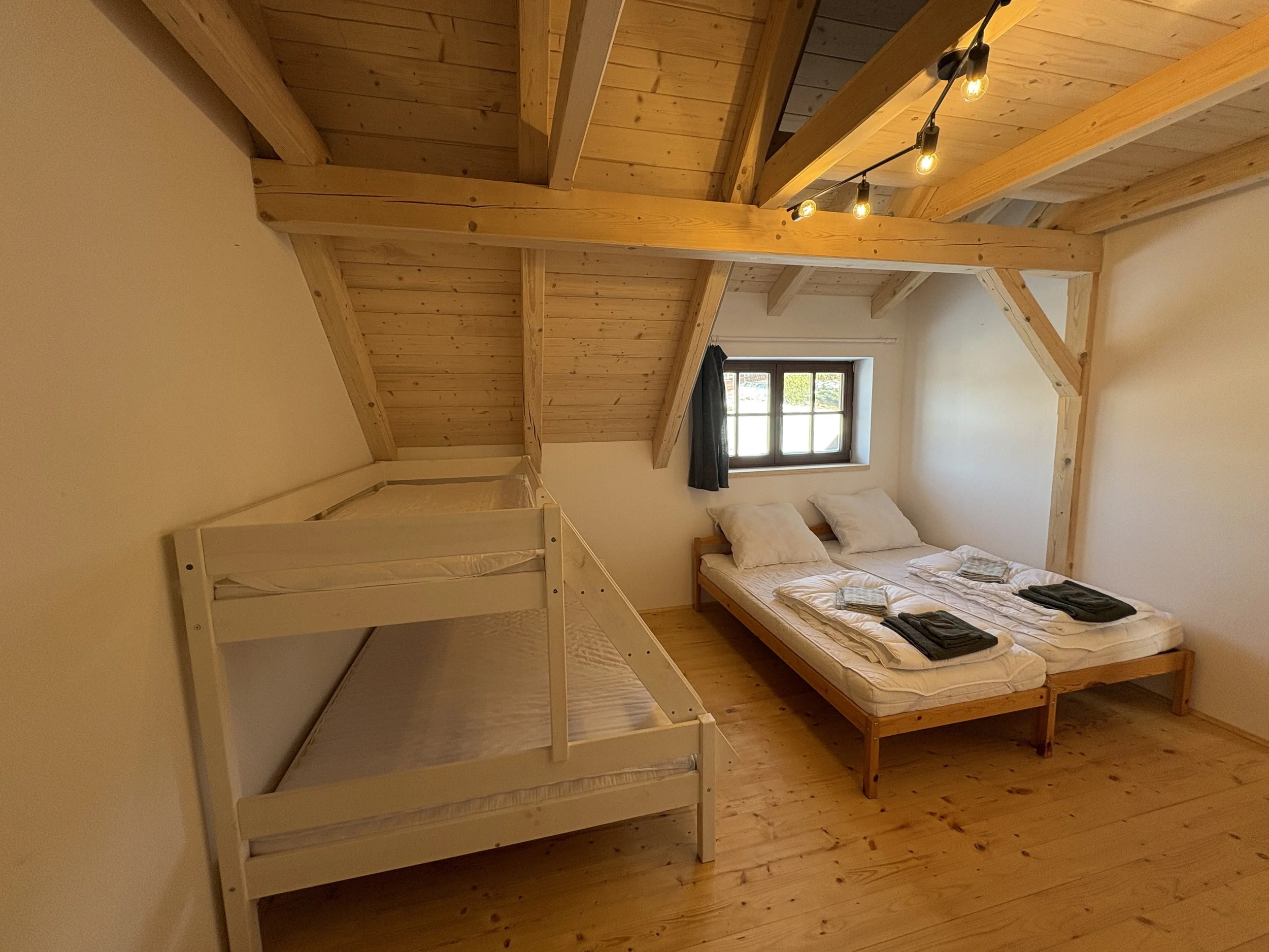 A cozy bedroom with wooden floors and sloped wooden ceiling beams. It features a small window, a double bed with two pillows and neatly folded towels, and a bunk bed with two mattresses, all in a minimalist style.