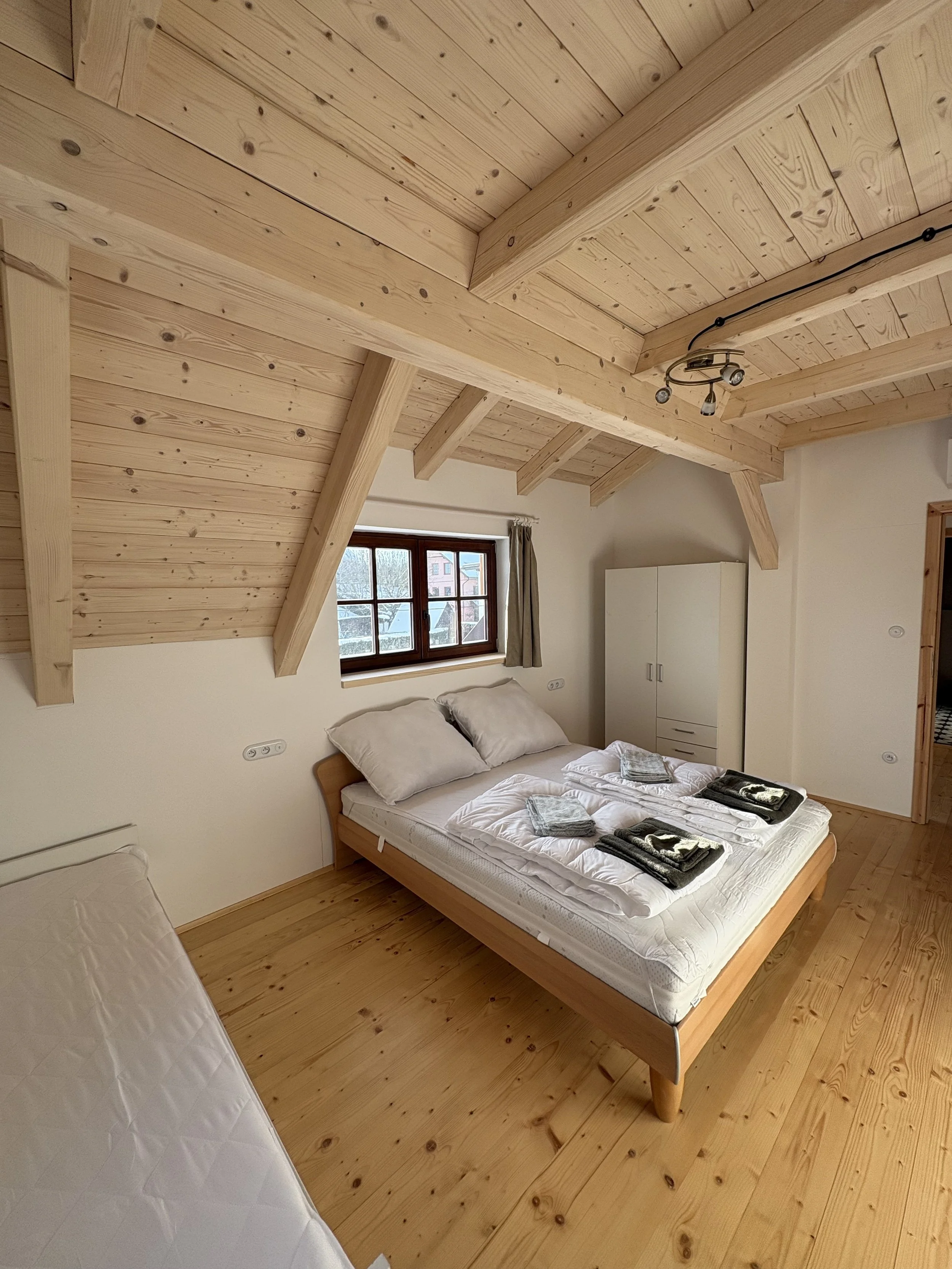 A cozy bedroom with wooden walls, ceiling, and floor. There is a large bed with white bedding and towels, a small window with a curtain, and a white wardrobe.