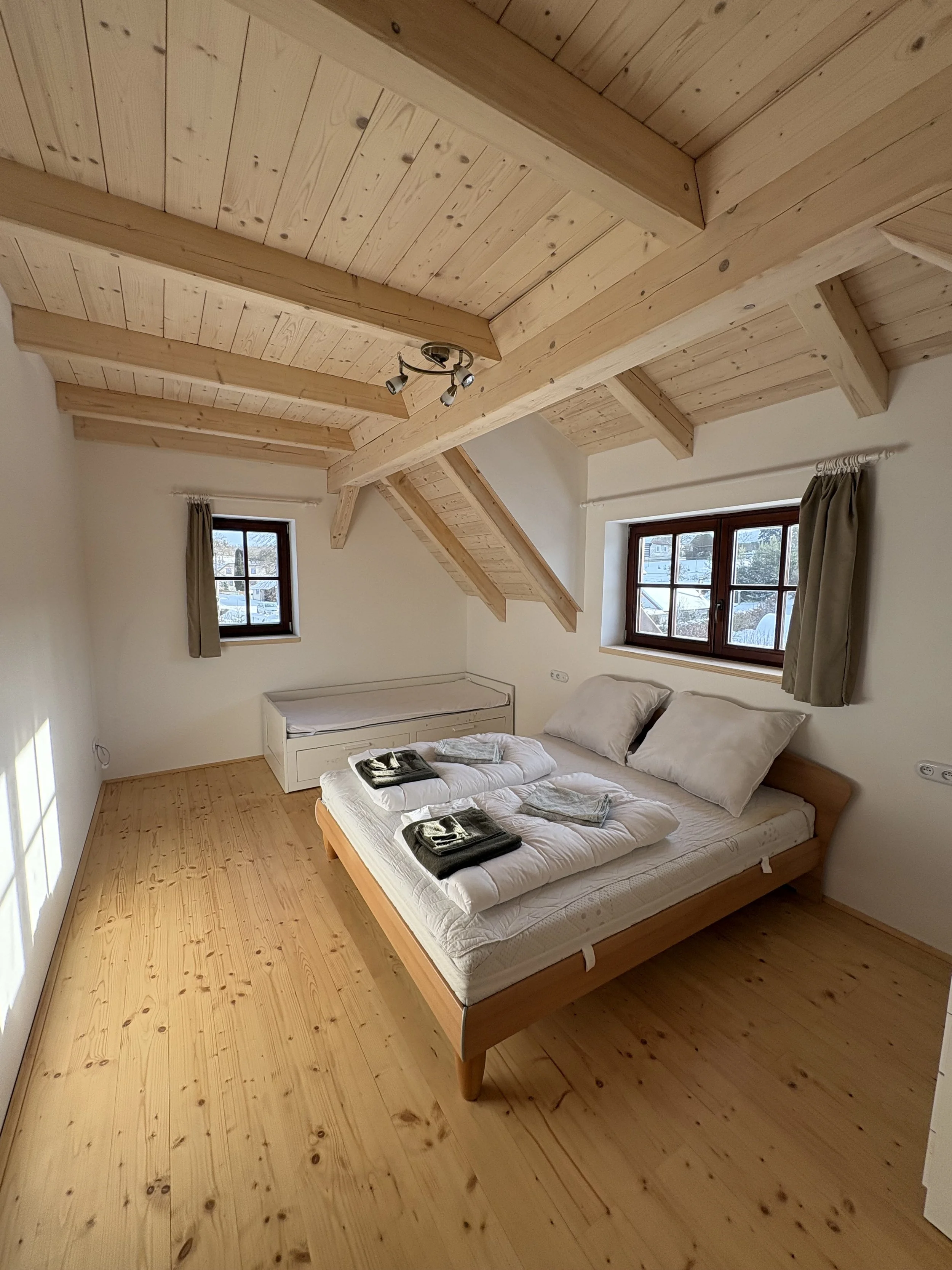 A spacious bedroom with wood-paneled ceiling and floor, featuring a wooden bed with white bedding, two pillows, and folded towels on the bed. There are two small windows with curtains, and a small bench or storage under the window at the back.