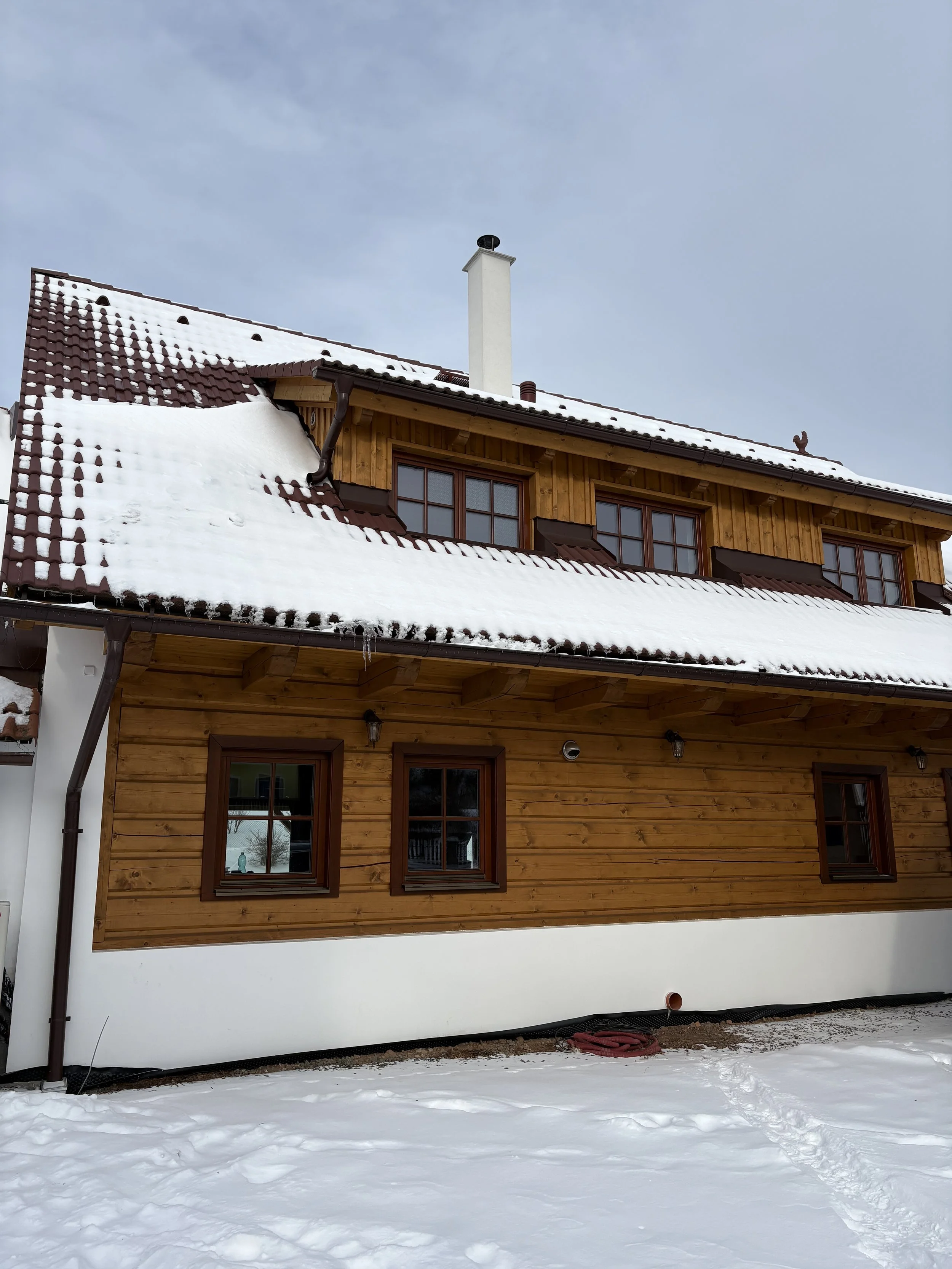 A wooden house with snow on its roof and around its foundation, four small windows on the ground floor, and five bigger windows on the upper floor, with a chimney and a cloudy sky in the background.