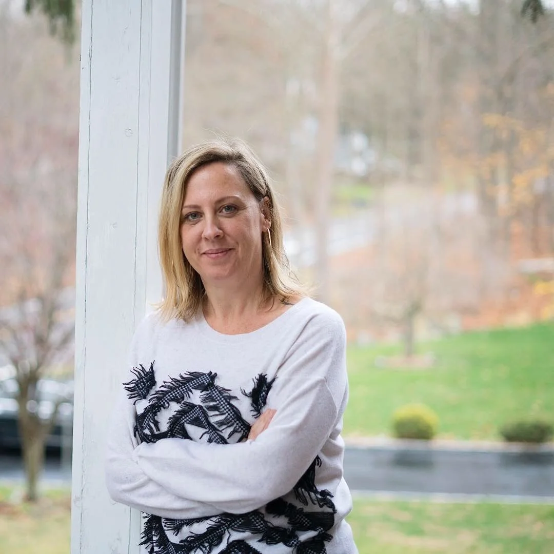 Photo of Jaime Harkin, a woman with medium length blonde hair, wearing a white long sleeve shirt with black feathers, leaning against a post with a blurred out background of a driveway with trees.