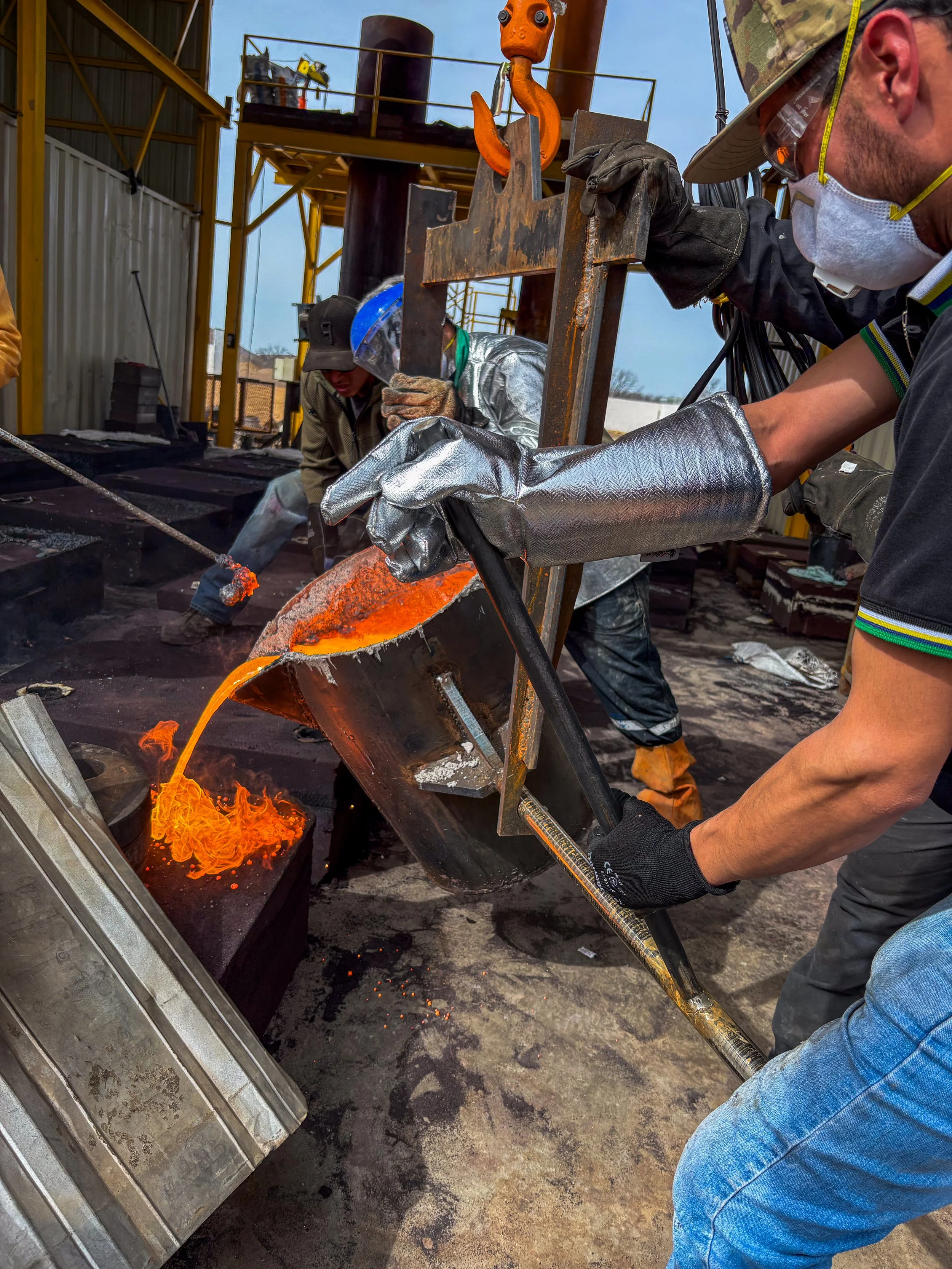 Trabajadores de metal fundiendo metal en un horno de fundición, vertiendo metal líquido en un molde, en un entorno de taller industrial.