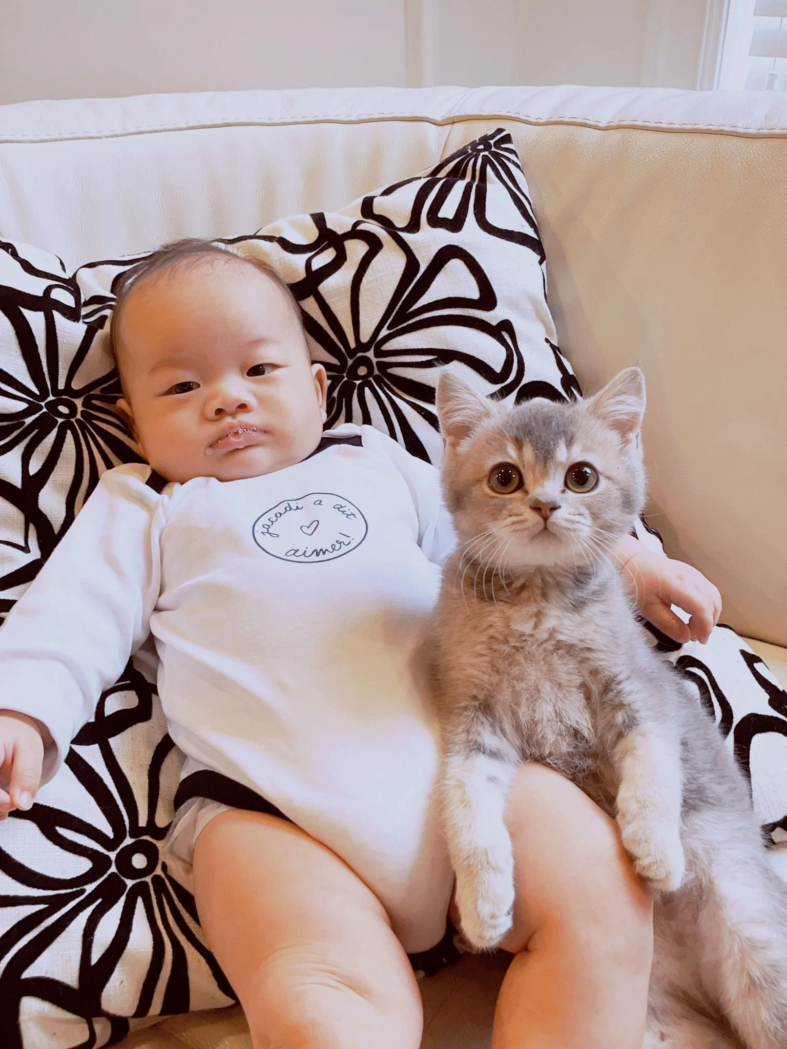 A baby lying on a black and white floral-patterned pillow on a beige sofa, holding a gray and cream kitten, with some milk or drool on their lips and shirt.