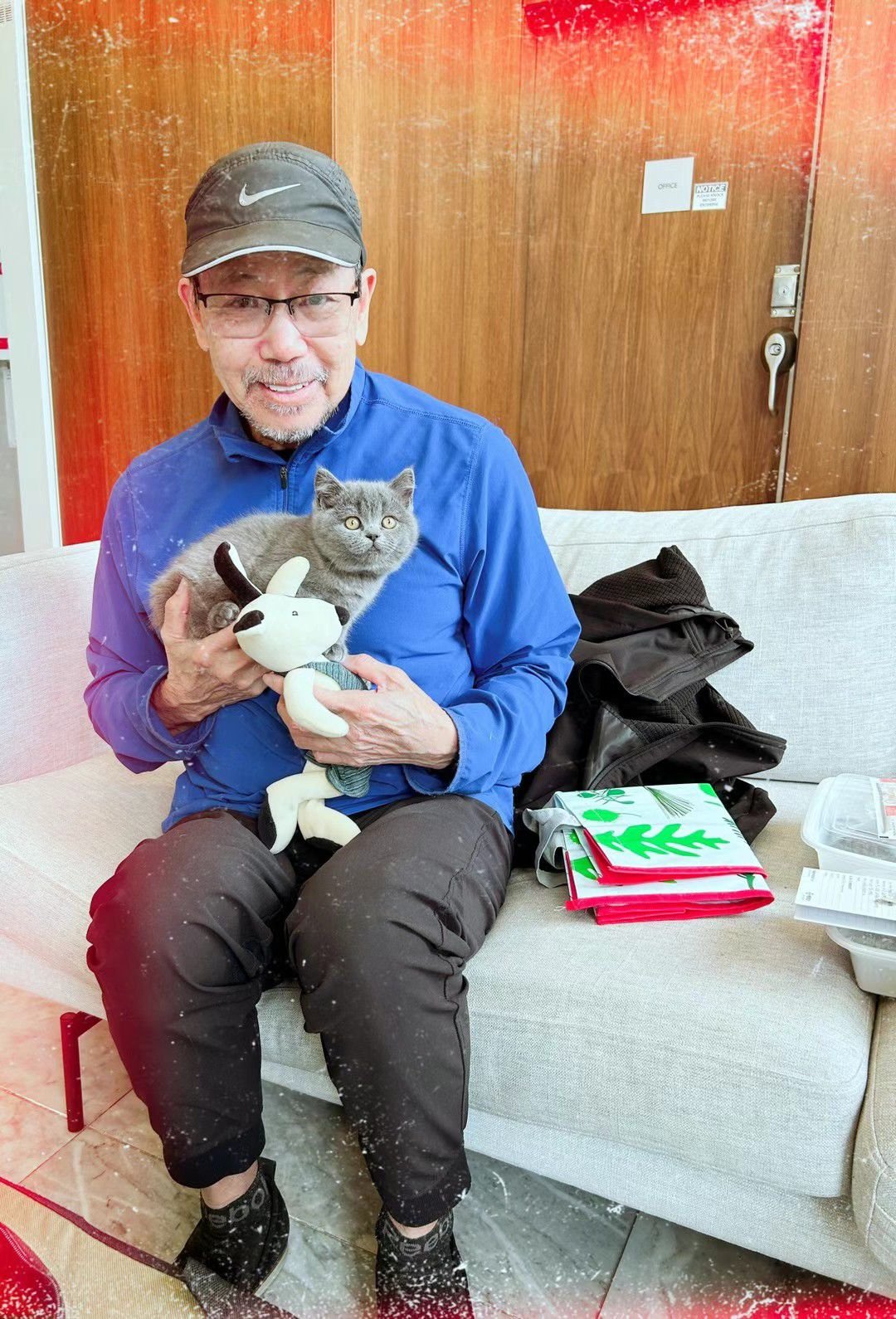 A man in a black cap, glasses, and blue jacket sitting on a beige sofa, holding a gray cat and a white plush toy. There are holiday gift bags and a black backpack on the sofa, with snow on the man's pants and shoes indicating a festive or winter setting.