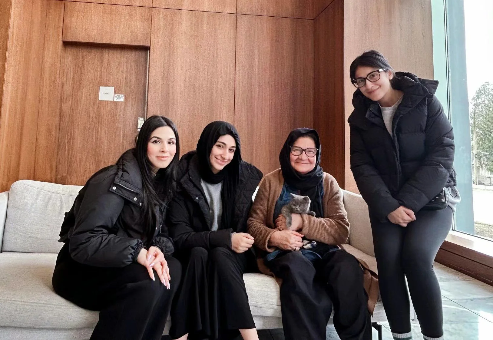 Four women sitting and standing on a beige sofa in a lobby or waiting area with wood-paneled walls and large windows, one woman holding a small gray kitten, all smiling at the camera.