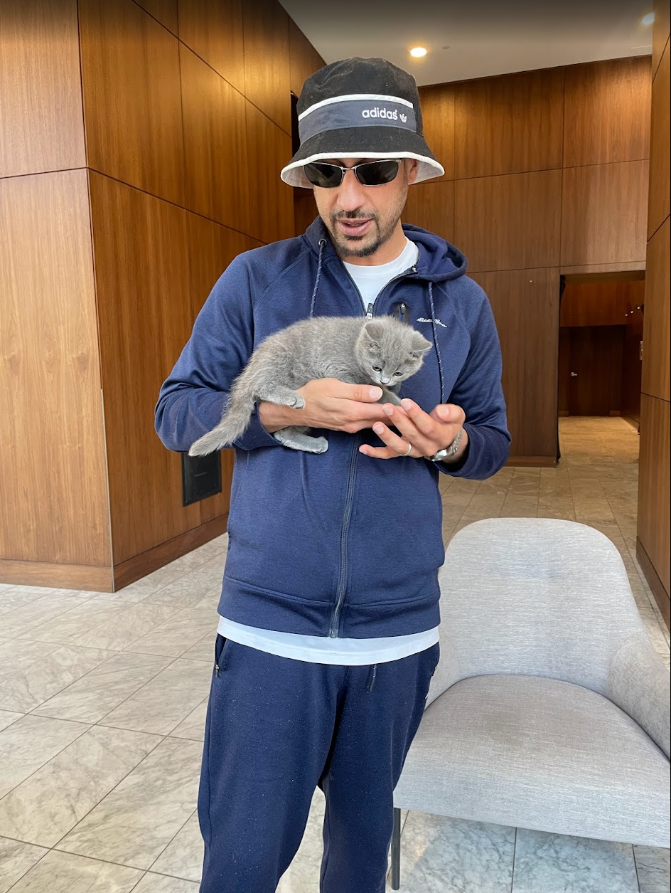 A man wearing sunglasses, a black bucket hat, a navy blue hoodie, and navy sweatpants holding a small gray kitten inside a room with wood-paneled walls and tiled flooring.