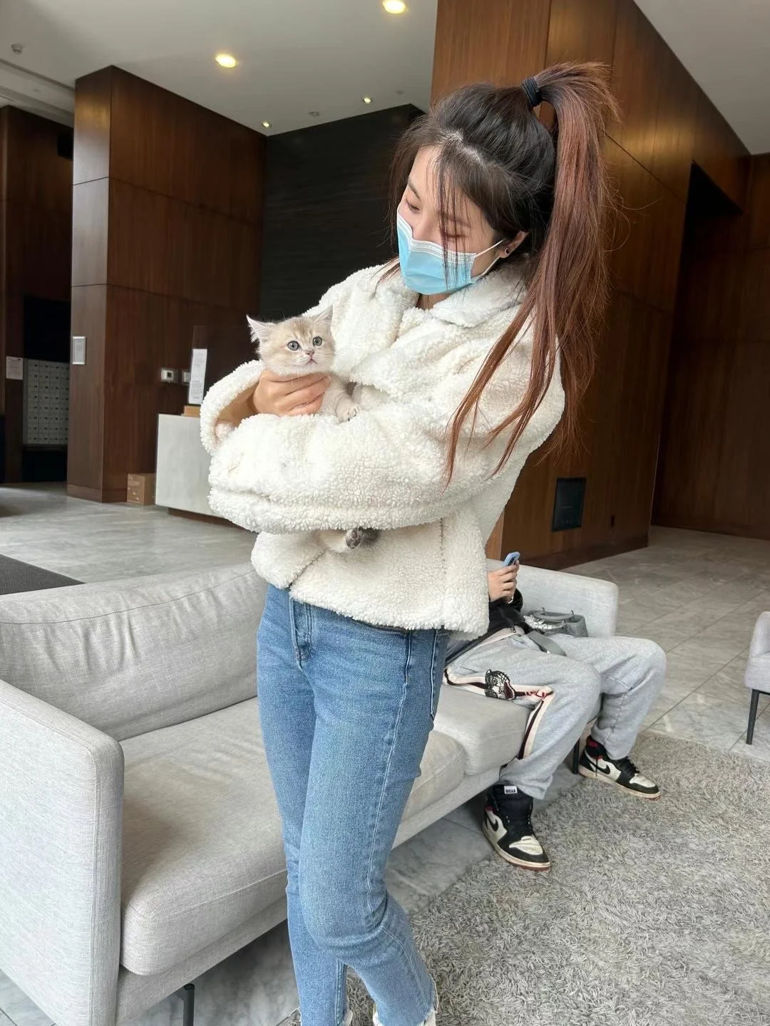 A woman in a blue face mask and a cream-colored fuzzy jacket holding a small kitten inside a modern living room.