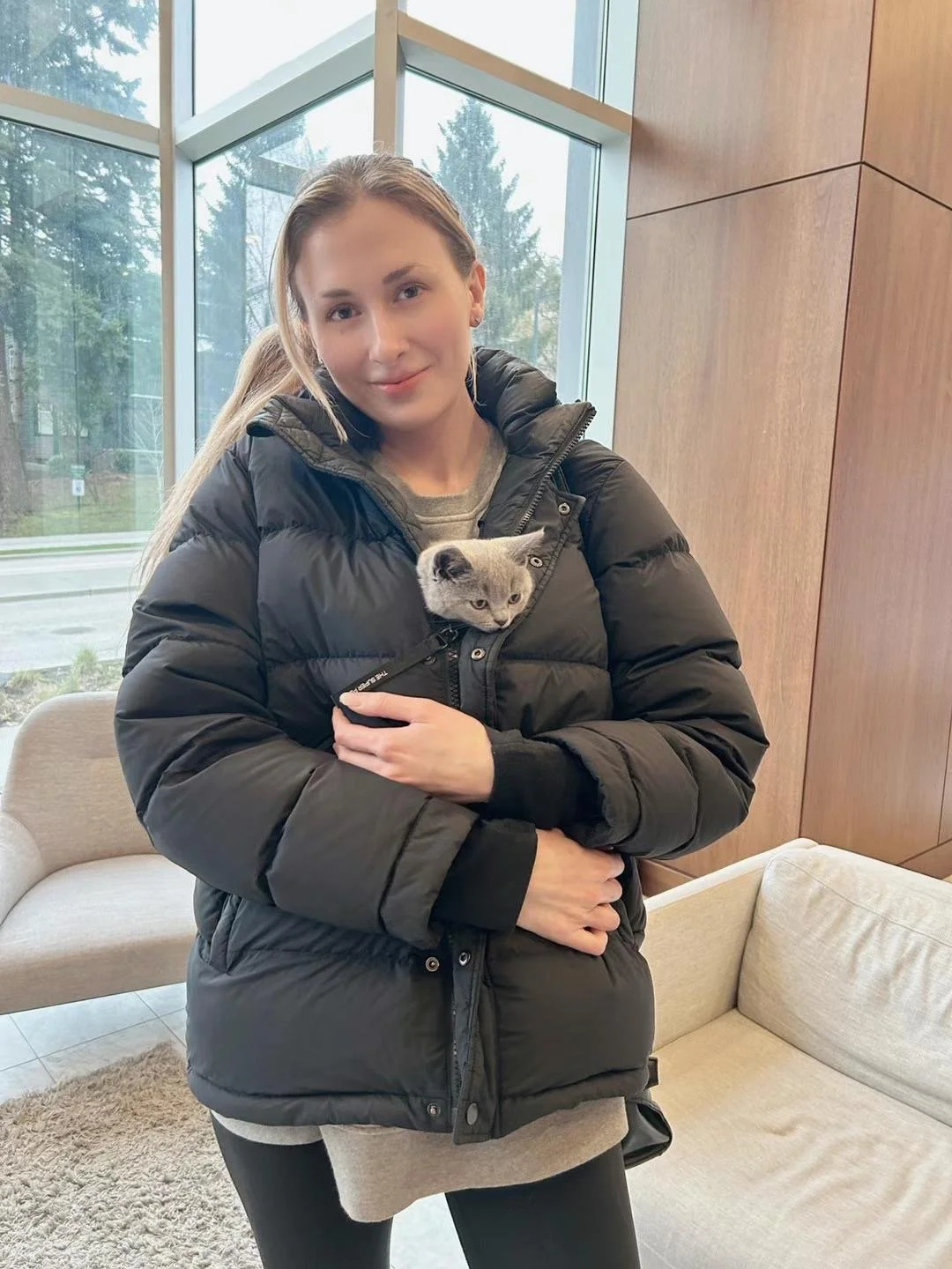 Young woman with a ponytail smiling while holding a gray kitten inside her black puffer jacket, sitting in a modern indoor space near large windows showing an outdoor scene with trees.