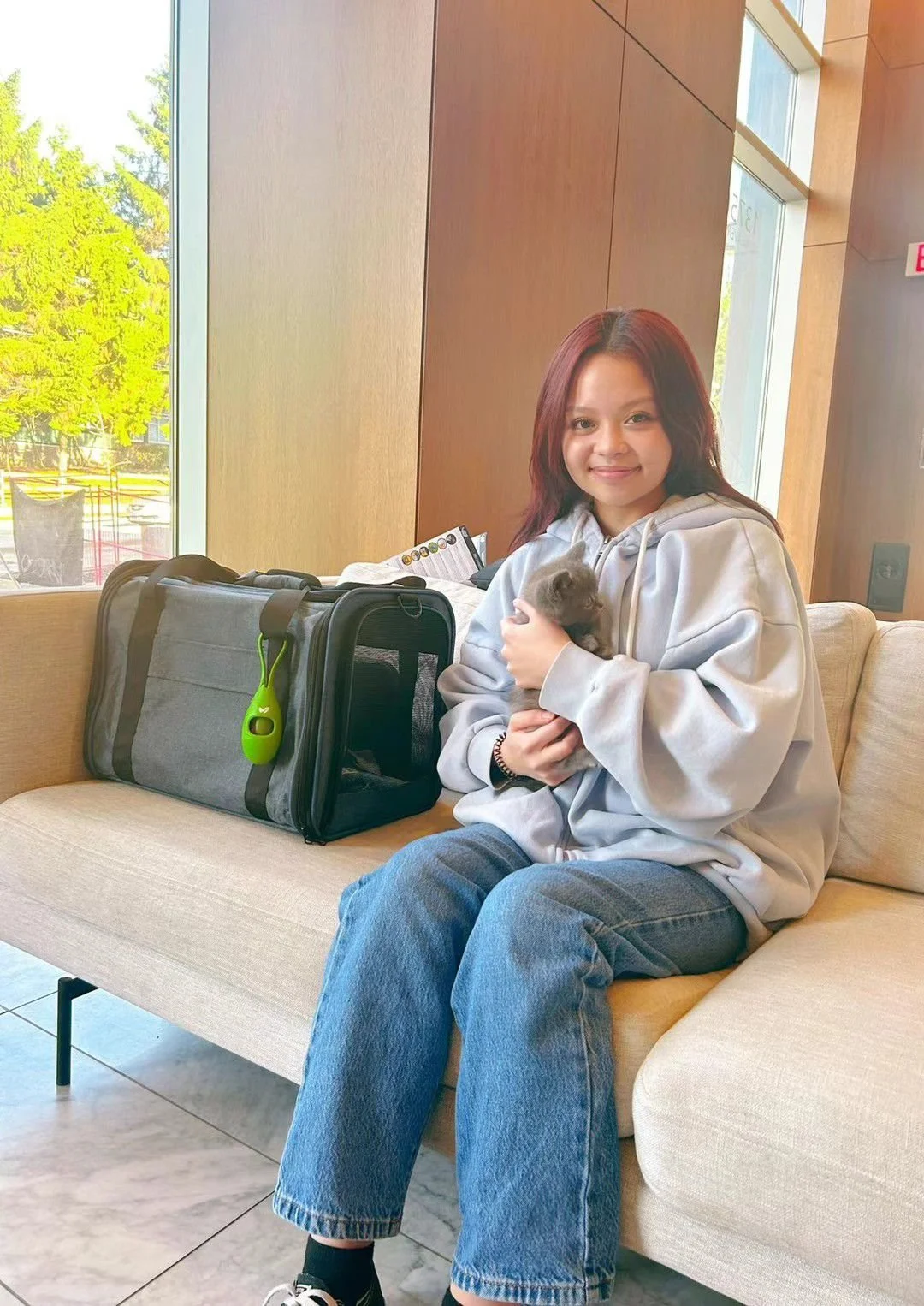 Young woman with auburn hair sitting on a beige sofa in a modern indoor space, holding a small gray kitten. Next to her is a large gray pet carrier with a green toy attached. Large windows behind her let in natural light, showing trees outside.