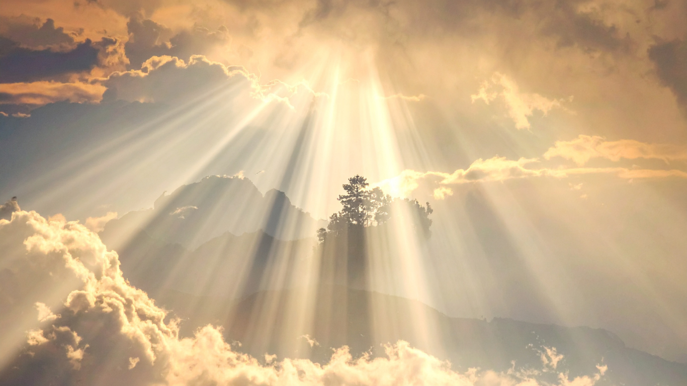 Sunlight rays shining through clouds over a mountainous landscape with trees on a hillside.