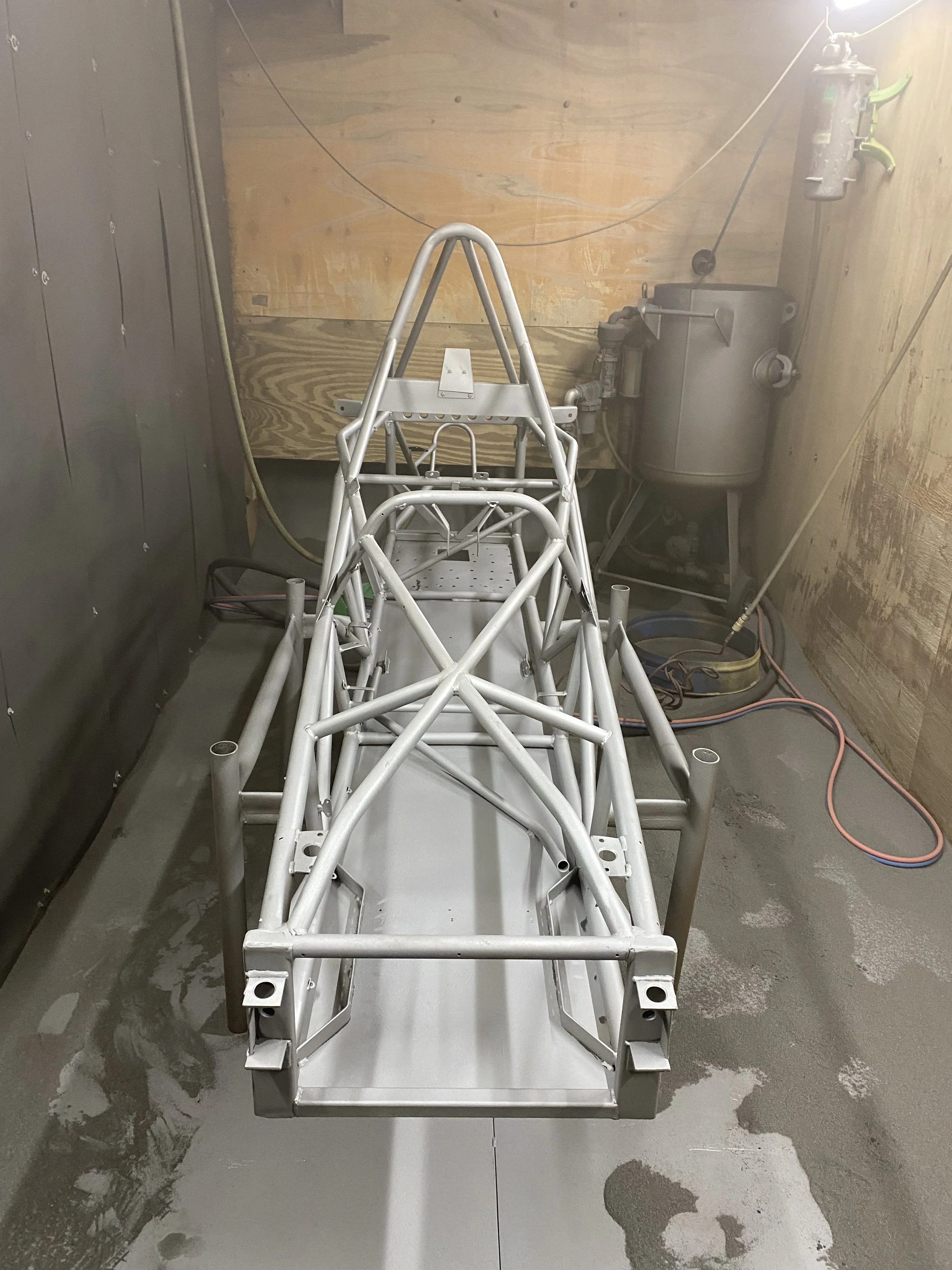 A metal frame chassis of a race car in a workshop or garage with wooden walls and concrete floor, alongside various tools and equipment.