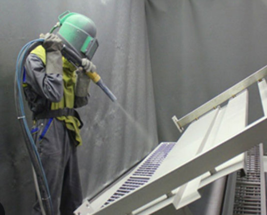 Person in protective suit and helmet operating a device with a nozzle in a gray industrial setting.