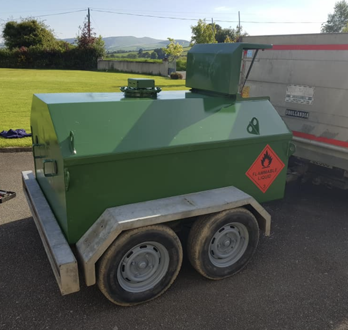 Green portable fuel tank on a two-wheel trailer, with a label indicating flammable liquid, parked outdoors on a paved surface with grass and trees in the background.