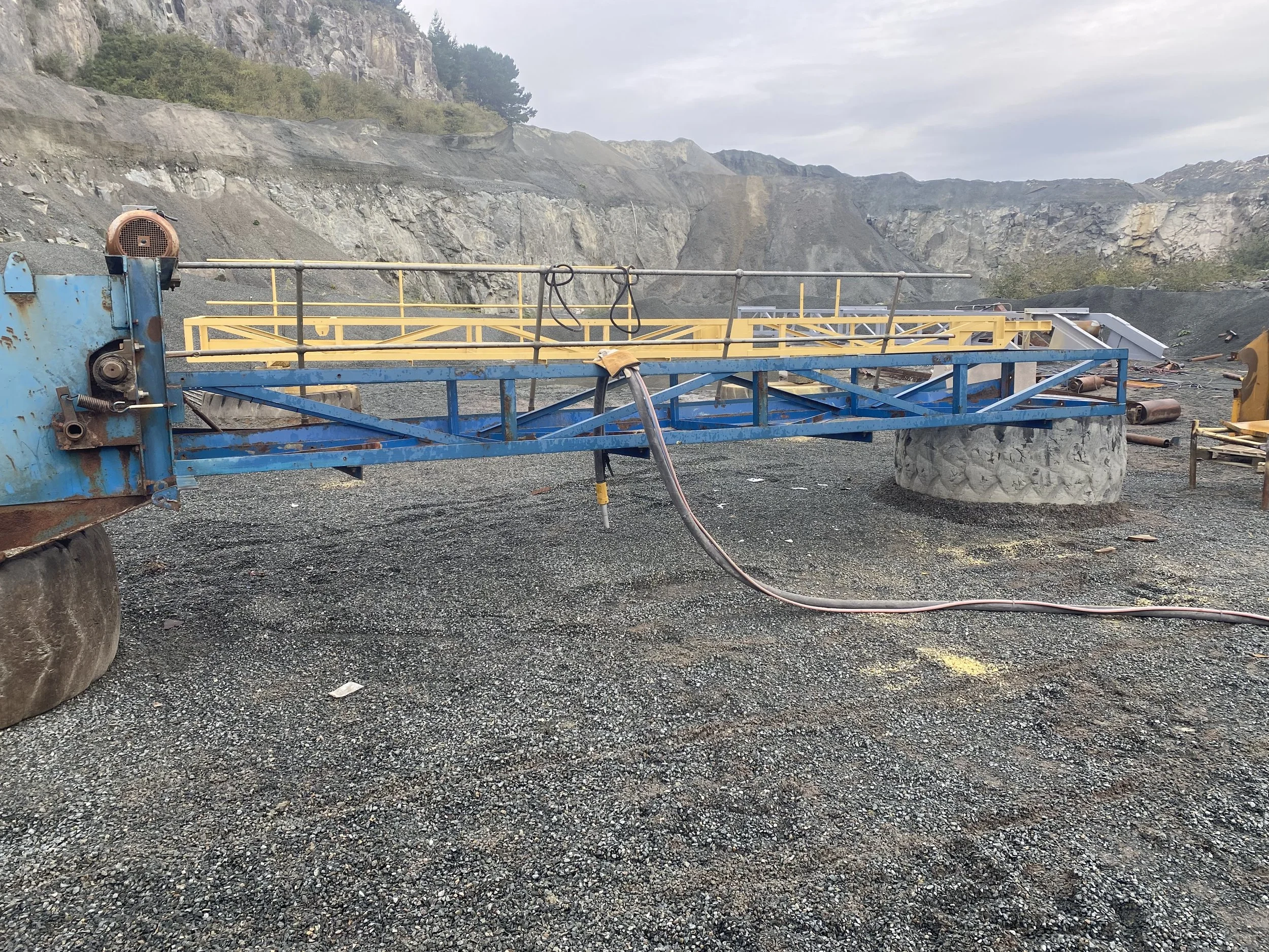 A blue and yellow construction crane with rust on parts, situated on gravel ground at a quarry, with rocky cliffs and industrial debris in the background.