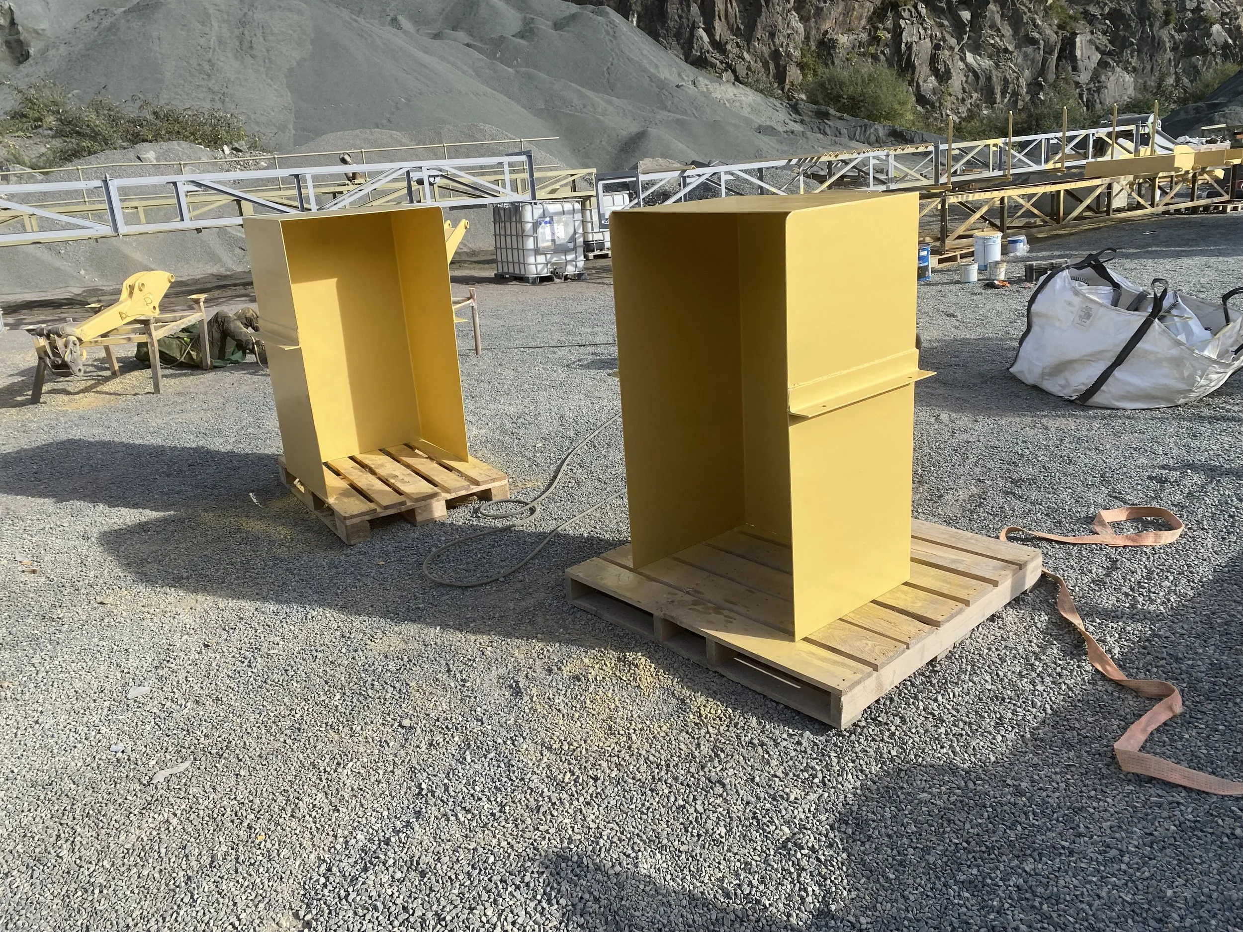 Two yellow metal structures on wooden pallets at a construction site with gravel ground, scaffolding, and rocky hills in the background.