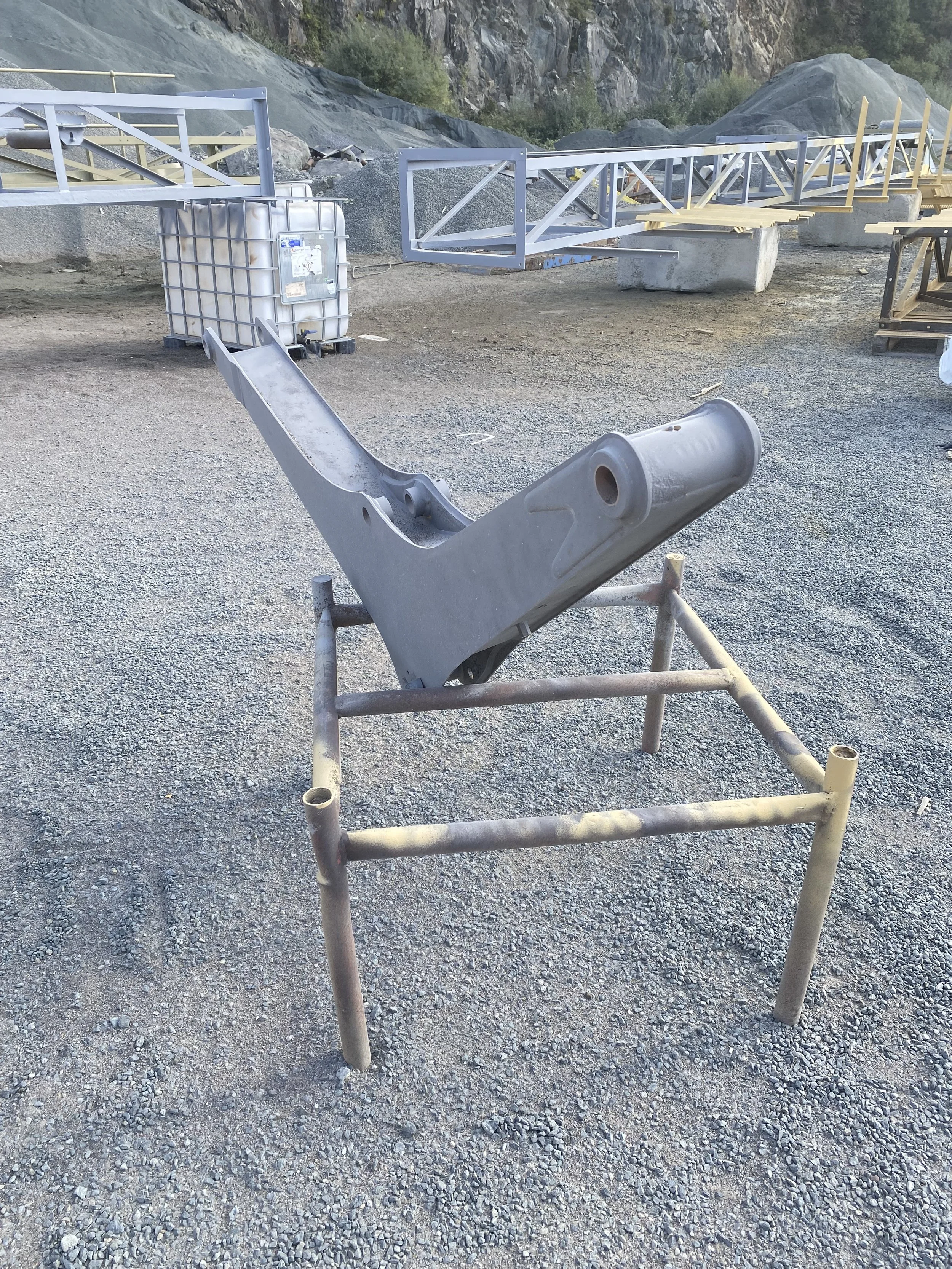 Metal frame and machinery components, with a large gray metal piece on a small metal stand in a gravel lot, with construction materials and rocky cliffs in the background.