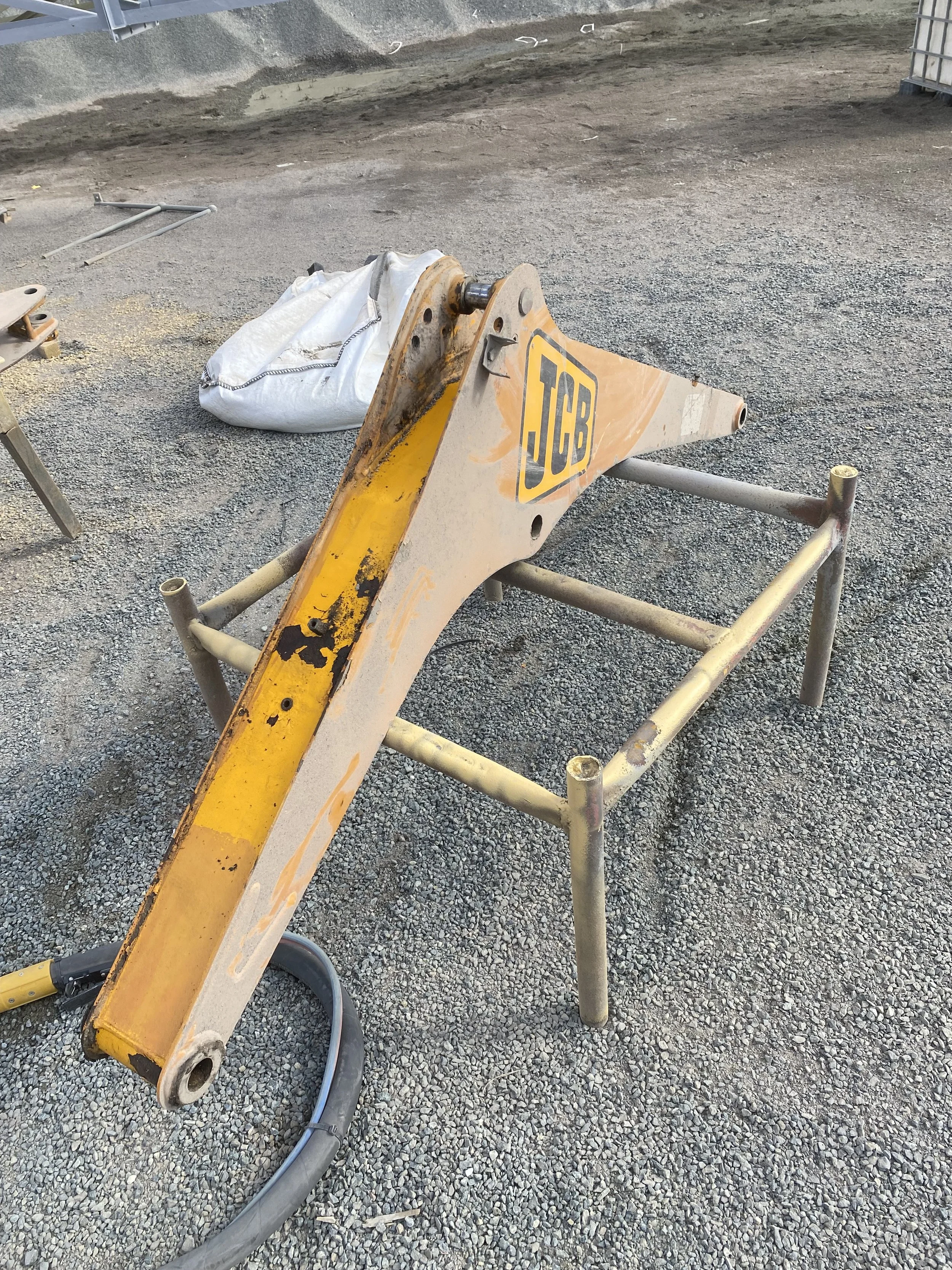 A JCB excavator arm resting on a metal stand outdoors on gravel, with a white bag and other construction equipment nearby.