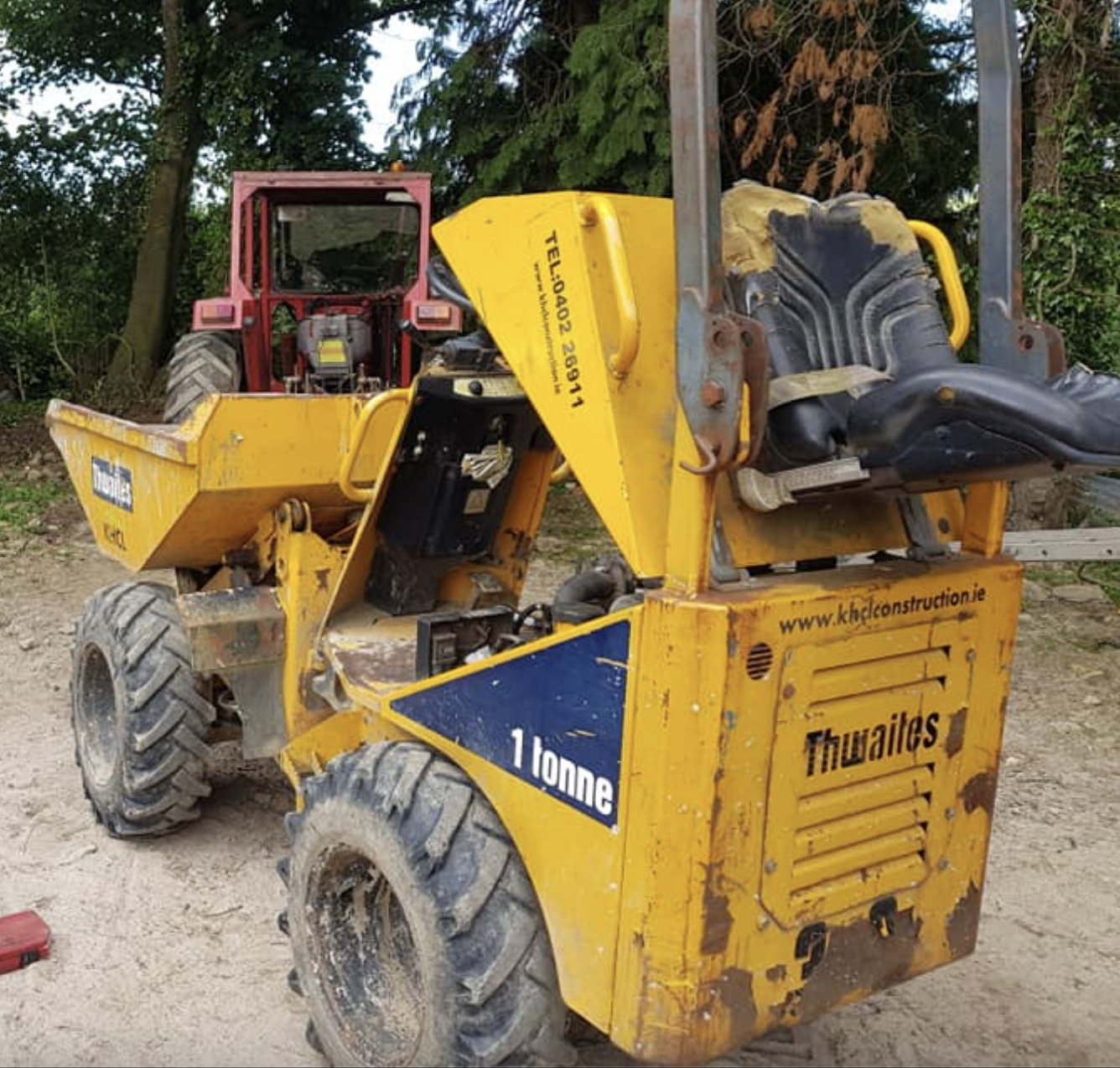 A yellow Thwaites dumper truck with a capacity of 1 tonne on construction site, with a red tractor in the background and trees surrounding the area.
