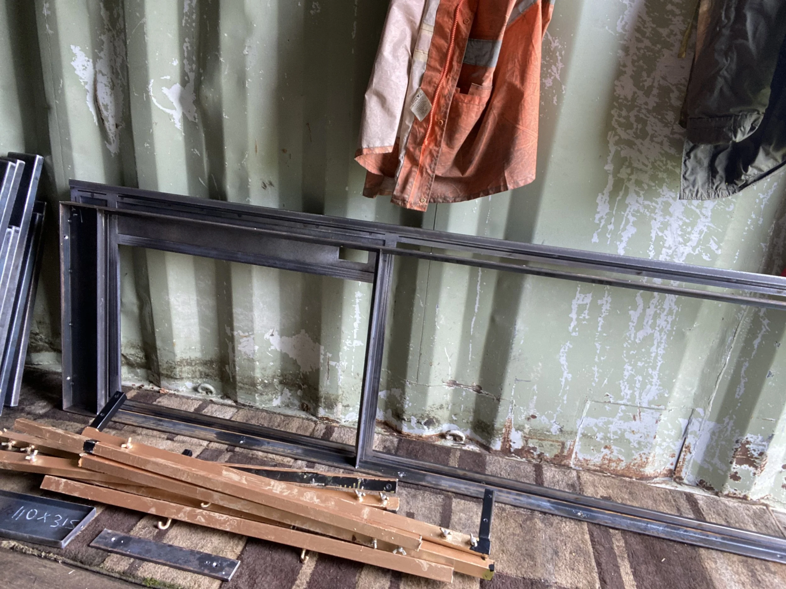 Empty metal and wooden door frames leaning against a green, worn wall with peeling paint, with jackets hanging on hooks above.