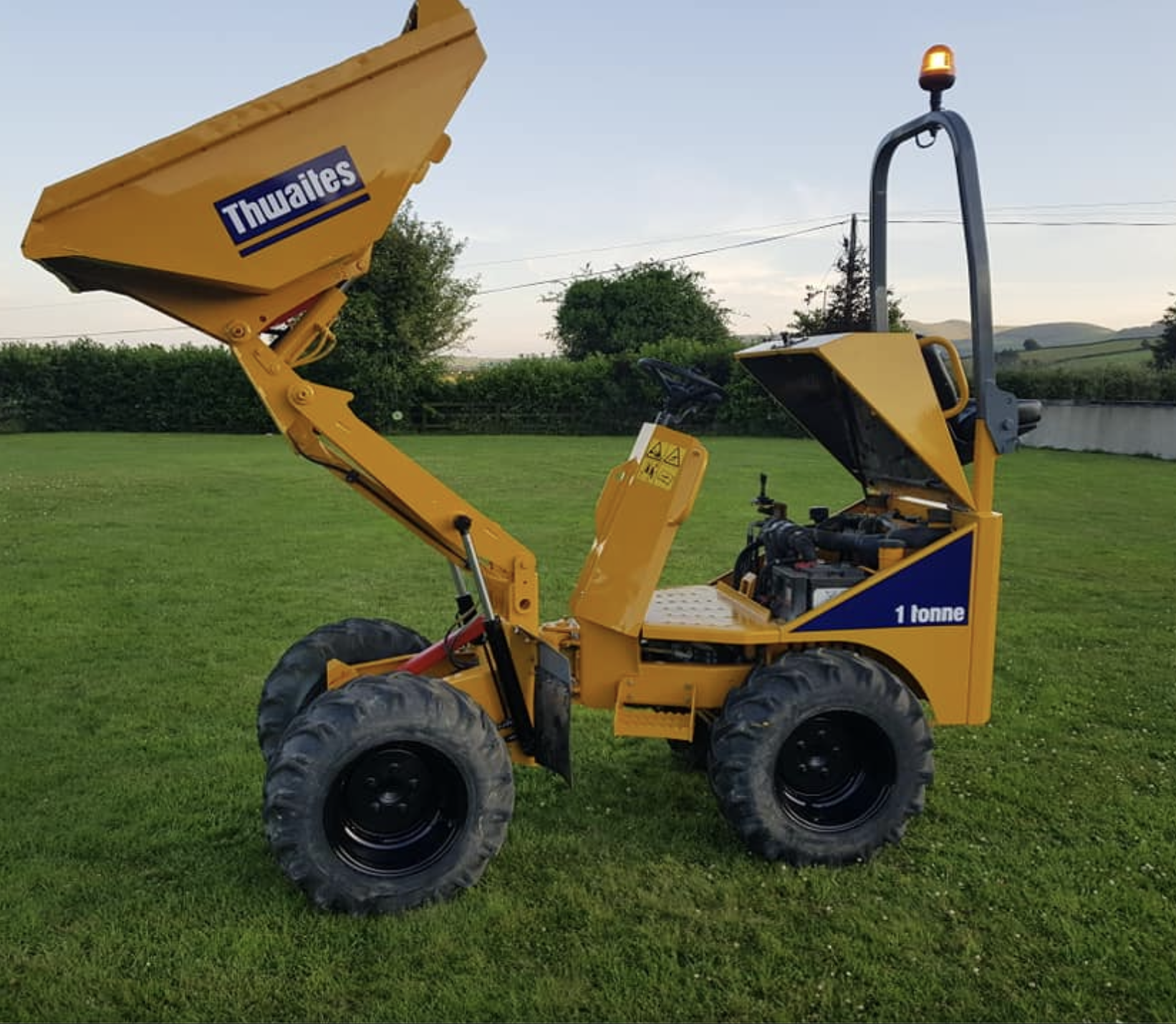 Small yellow piece of construction equipment with a lifted front loading bucket and a small enclosed cab, parked on grass in a rural area.