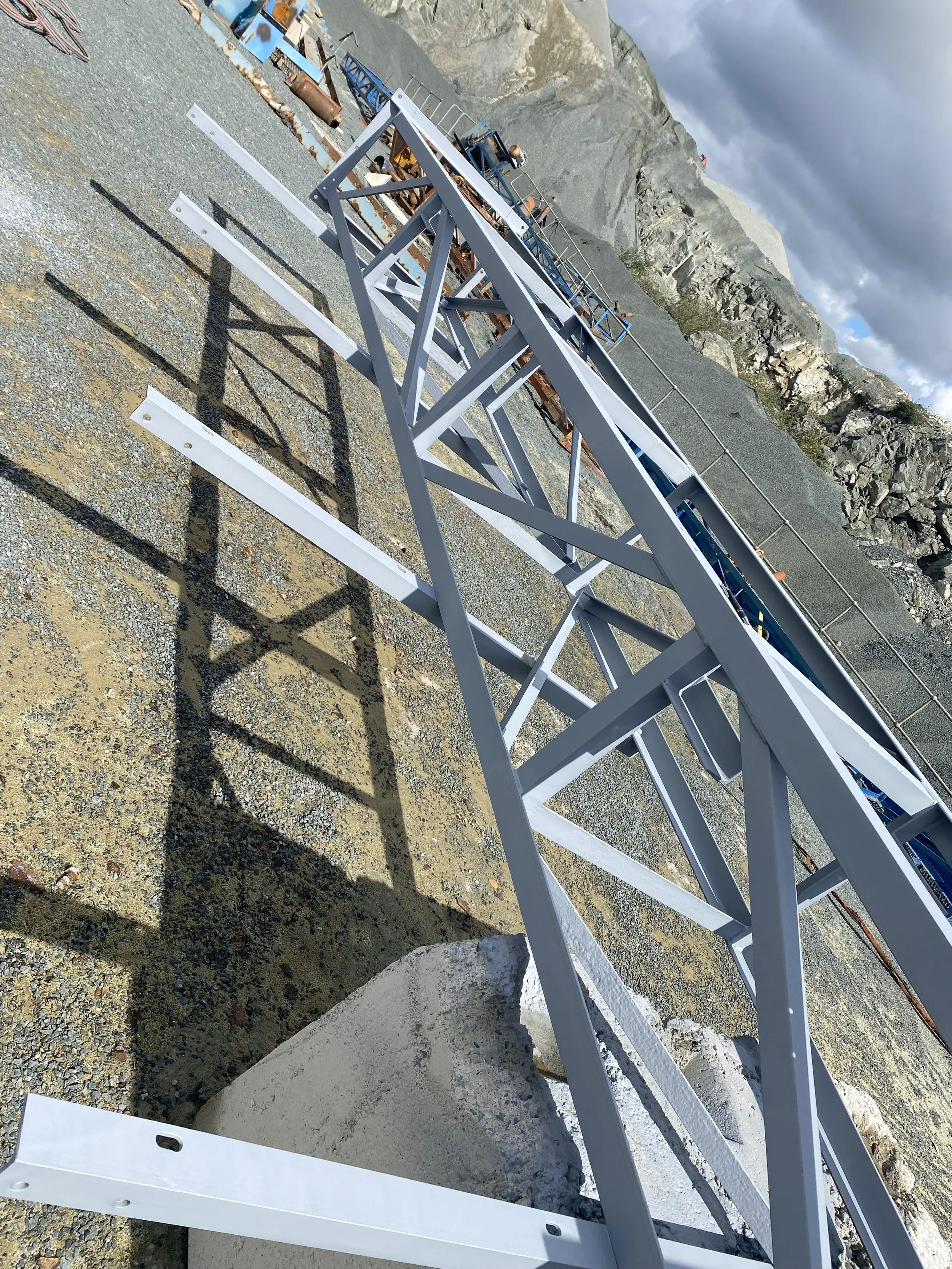 A long metal framework structure, supported by a concrete block, is situated on a gravel surface in an outdoor area with rocky hills in the background.