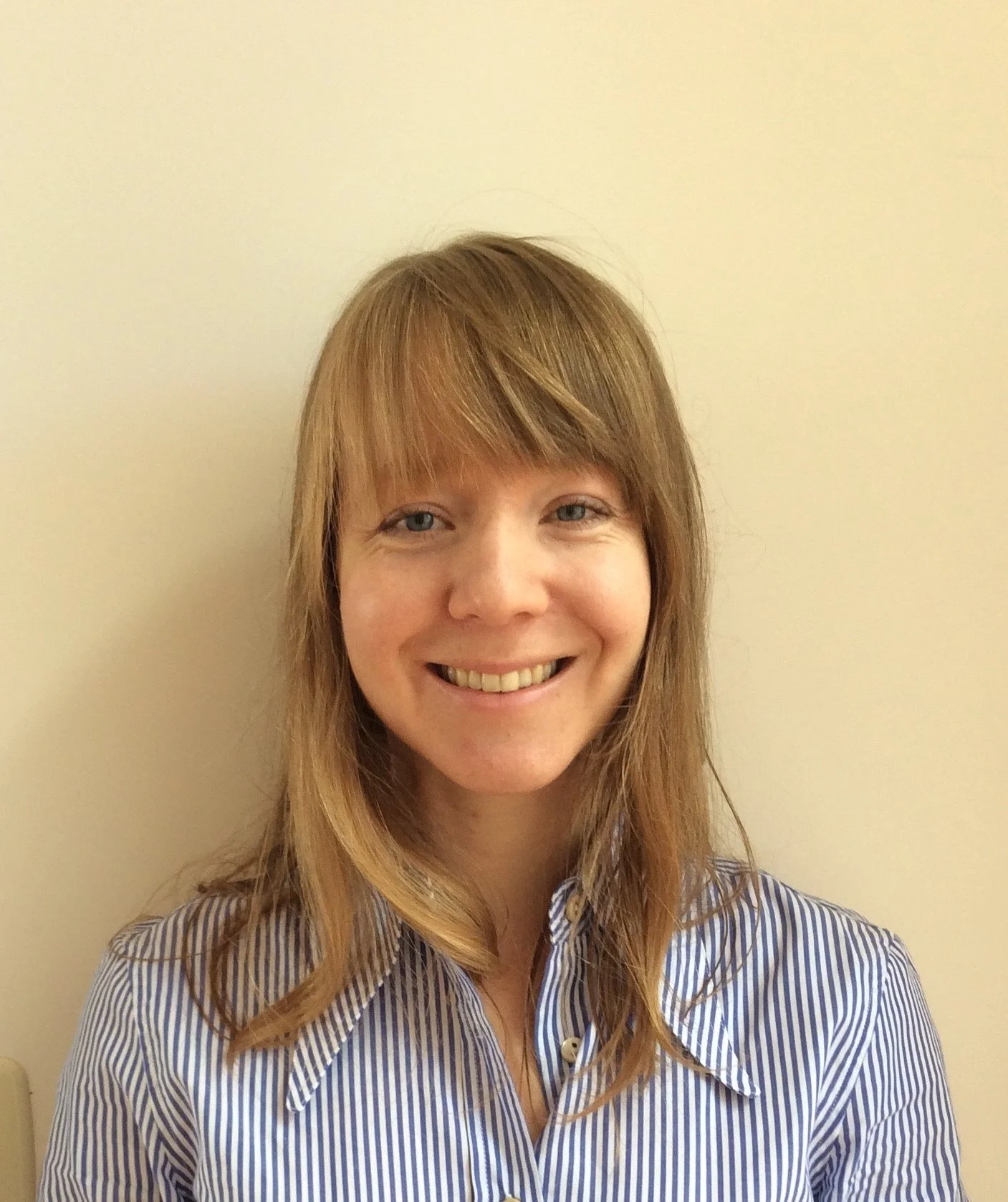 A woman with reddish-brown hair, blue eyes, smiling, wearing a blue and white striped button-up shirt, standing against a plain light-colored wall.