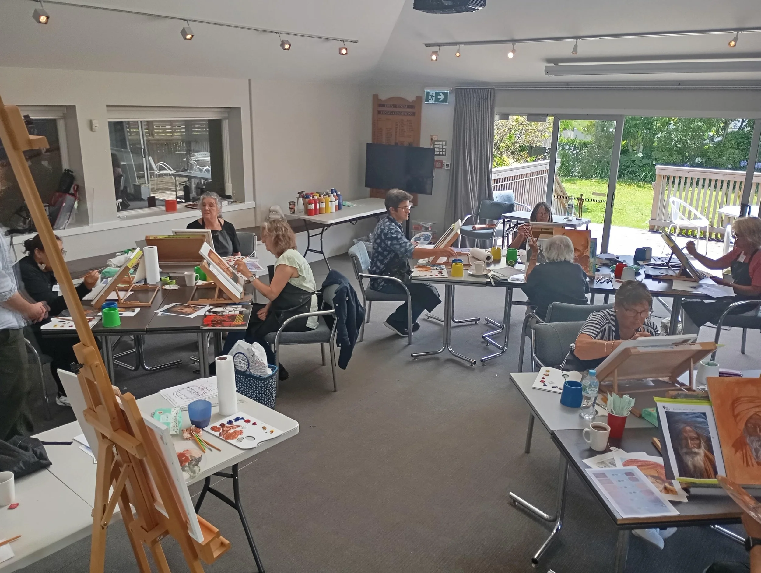 A group of people participating in a painting class in a bright room with large glass doors leading outside to a grassy yard.