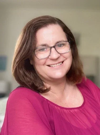 A woman with shoulder-length brown hair, glasses, and a bright smile, wearing a pink top, standing indoors against a neutral background.