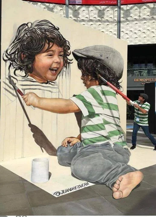 A street art mural of a young boy with curly hair wearing a striped shirt and gray cap, smiling and holding a paintbrush. A person standing nearby appears to be painting the wall to make the mural look like the boy is the artist, creating a playful optical illusion.