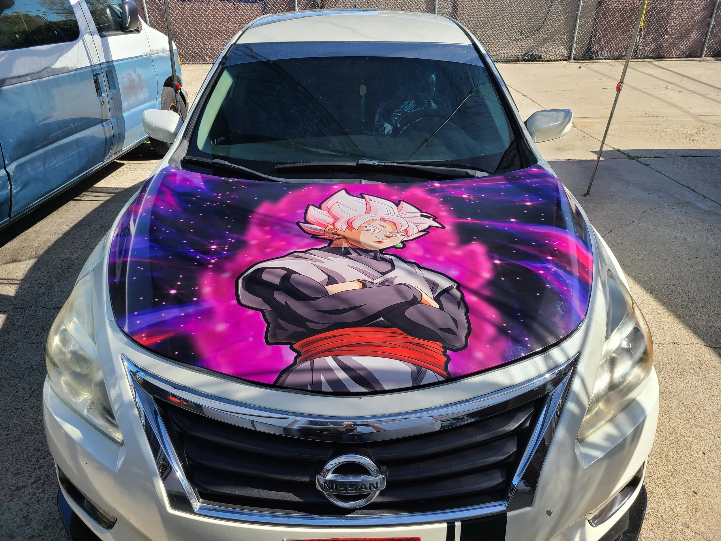 A silver Nissan car with a large graphic of Goku Black from Dragon Ball Super on the hood, with crossed arms and pink and purple cosmic background.