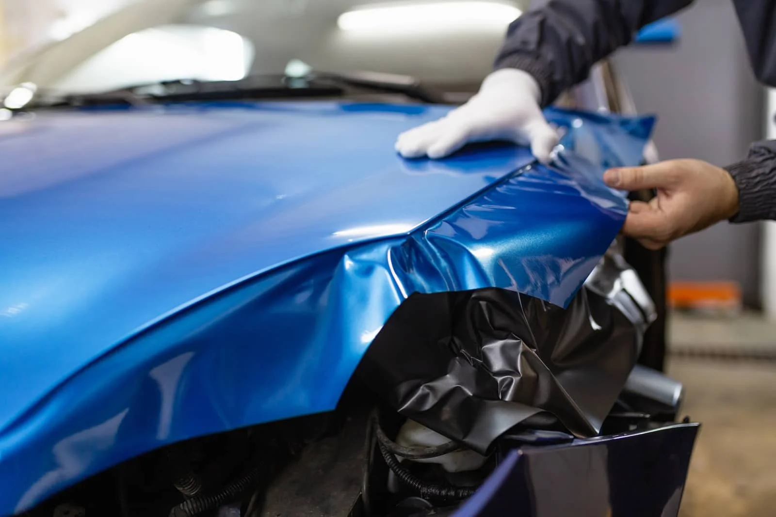 A person is applying blue vehicle wrap film to a car body, using a squeegee. The person is wearing gloves and a dark jacket.