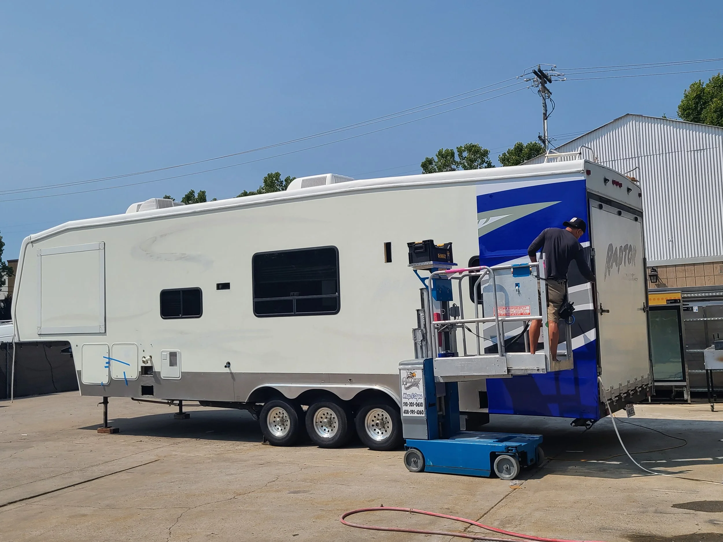 A person working on the back of a white and blue RV, standing on a small lift platform with tools nearby, outside on a paved lot.