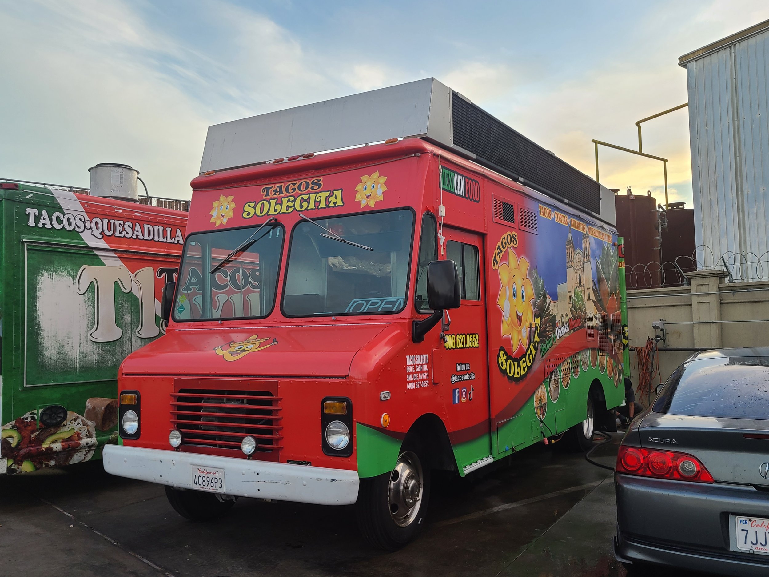 Red food truck with graphics of a smiling sun mascot and a collage of burritos, tacos, and other Mexican dishes, parked at an outdoor lot during sunset.
