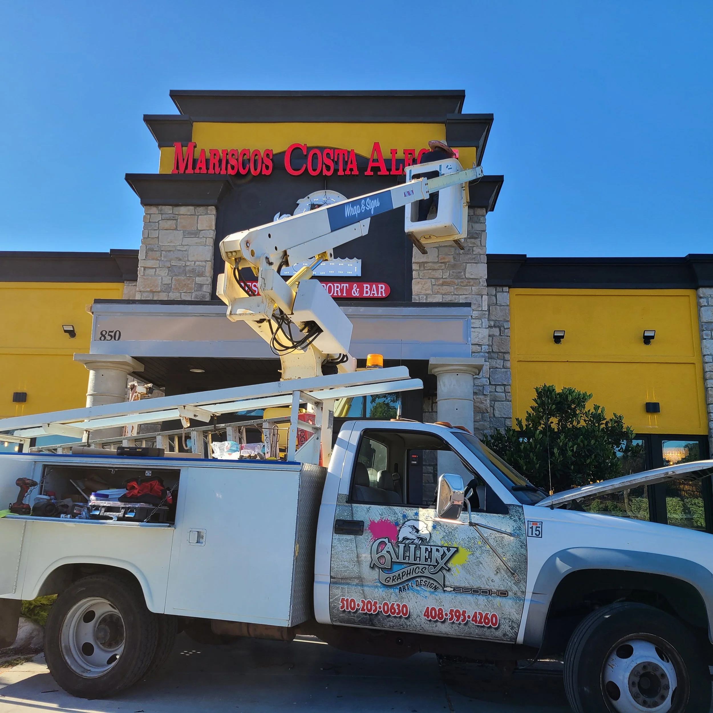 A utility truck with a hydraulic lift is parked outside a restaurant called Mariscos Costa Alegría. The truck has company branding and contact numbers on the side.
