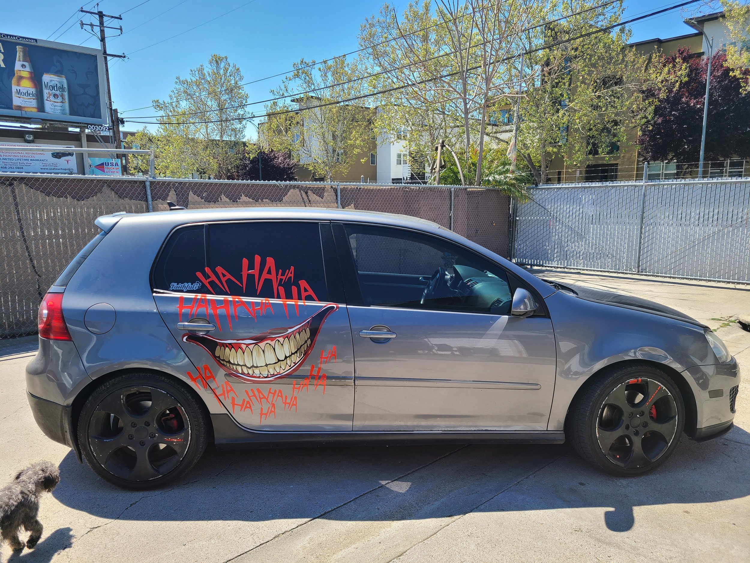 A silver hatchback car with a large graphic of a wide, toothy, smiling mouth and the words "HAHA" in red graffiti-style text on the side window. The car is parked on a concrete surface, with a small dog and a fence in the background.