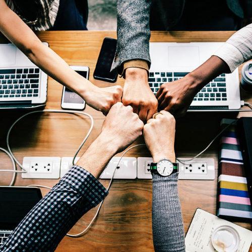 Six people join their fists together over a wooden table with laptops, phones, and office supplies.