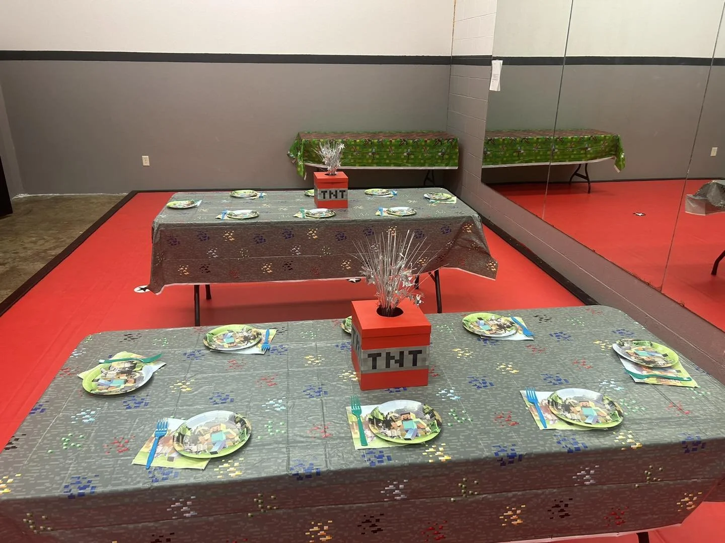 Party table decorated with themed plates and colorful napkins, featuring a TNT box centerpiece with metallic silver confetti and a green tablecloth in a room with red carpet and mirrored wall.