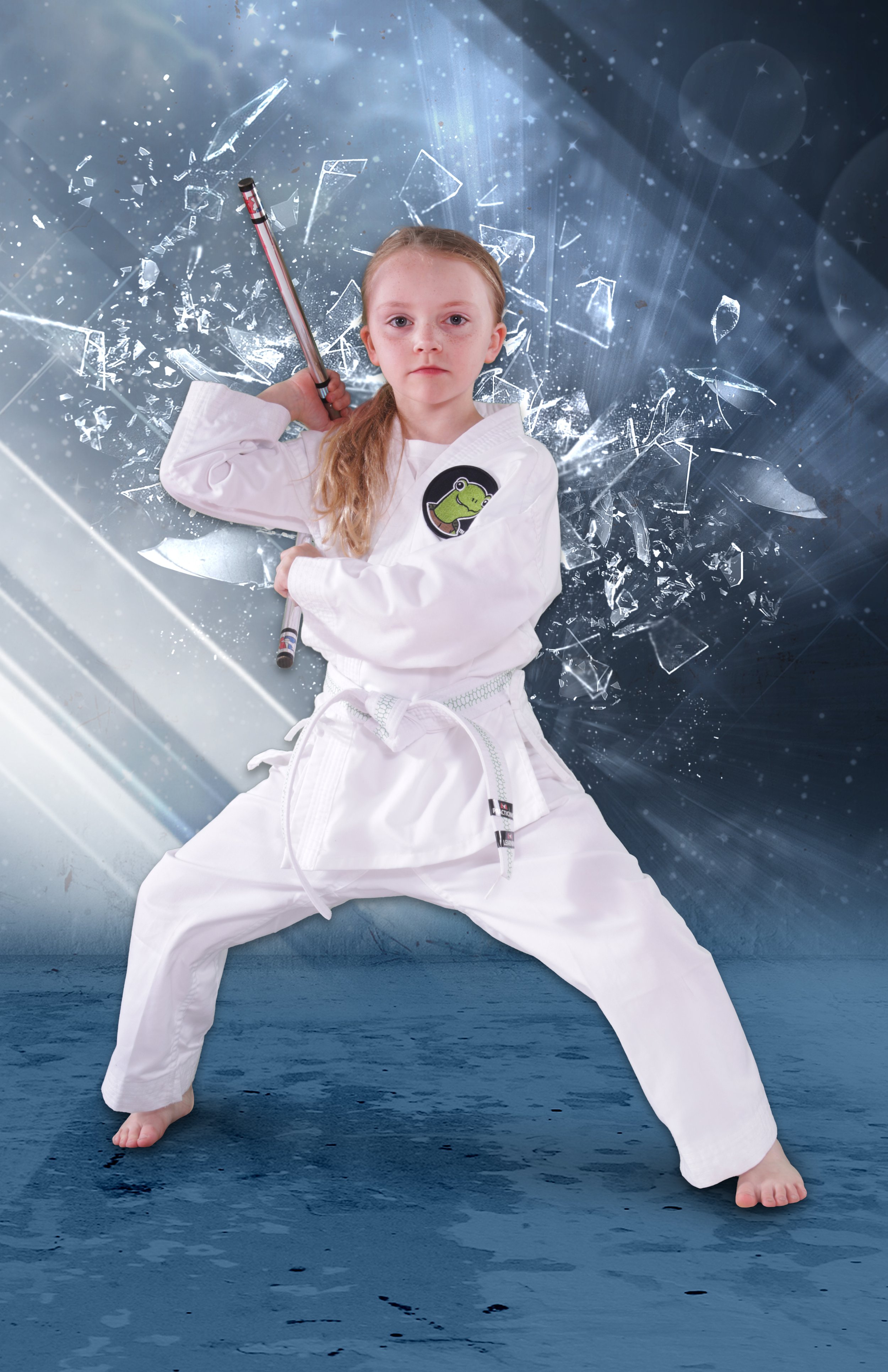 Young girl in a martial arts uniform with a frog patch, holding a stick, striking a fighting pose in front of a background with shattered glass and digital effects.
