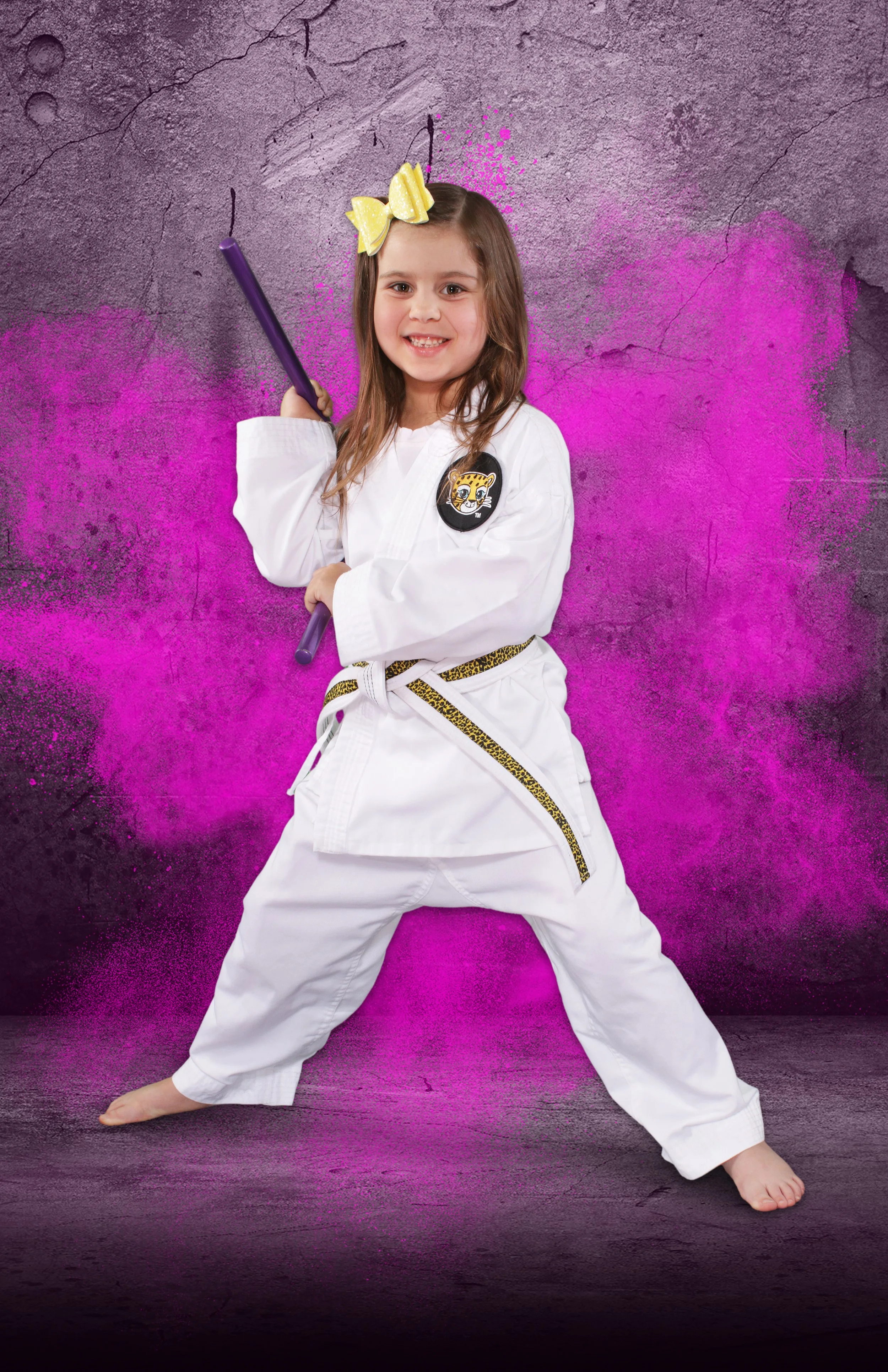 a photo of a young girl in martial arts uniform and a hair bow, holding nunchuku and smiling.